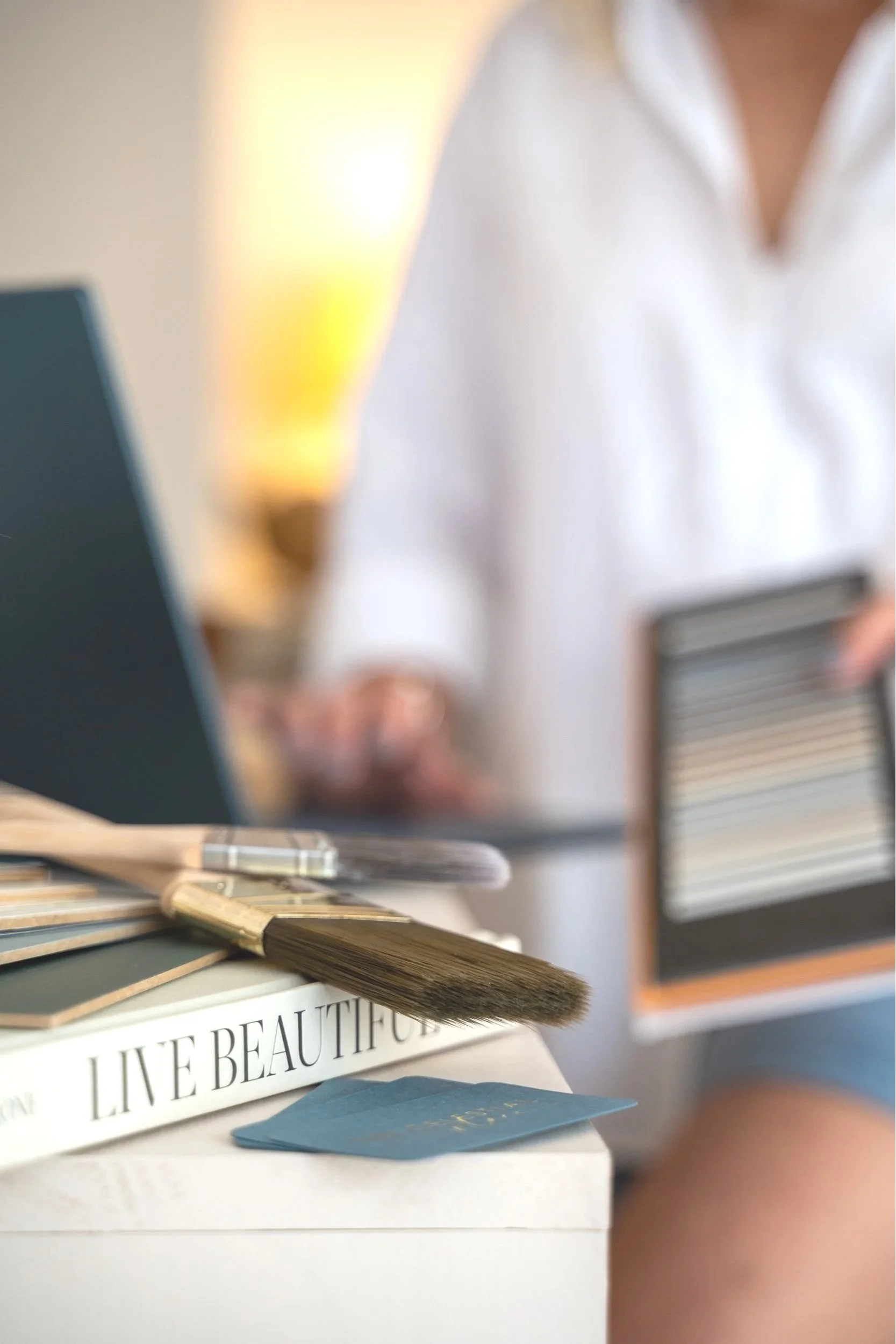 Close-up of a desk with a book titled 'Live Beautiful,' some color swatches, and a blue card, with a person in a white shirt in the background.