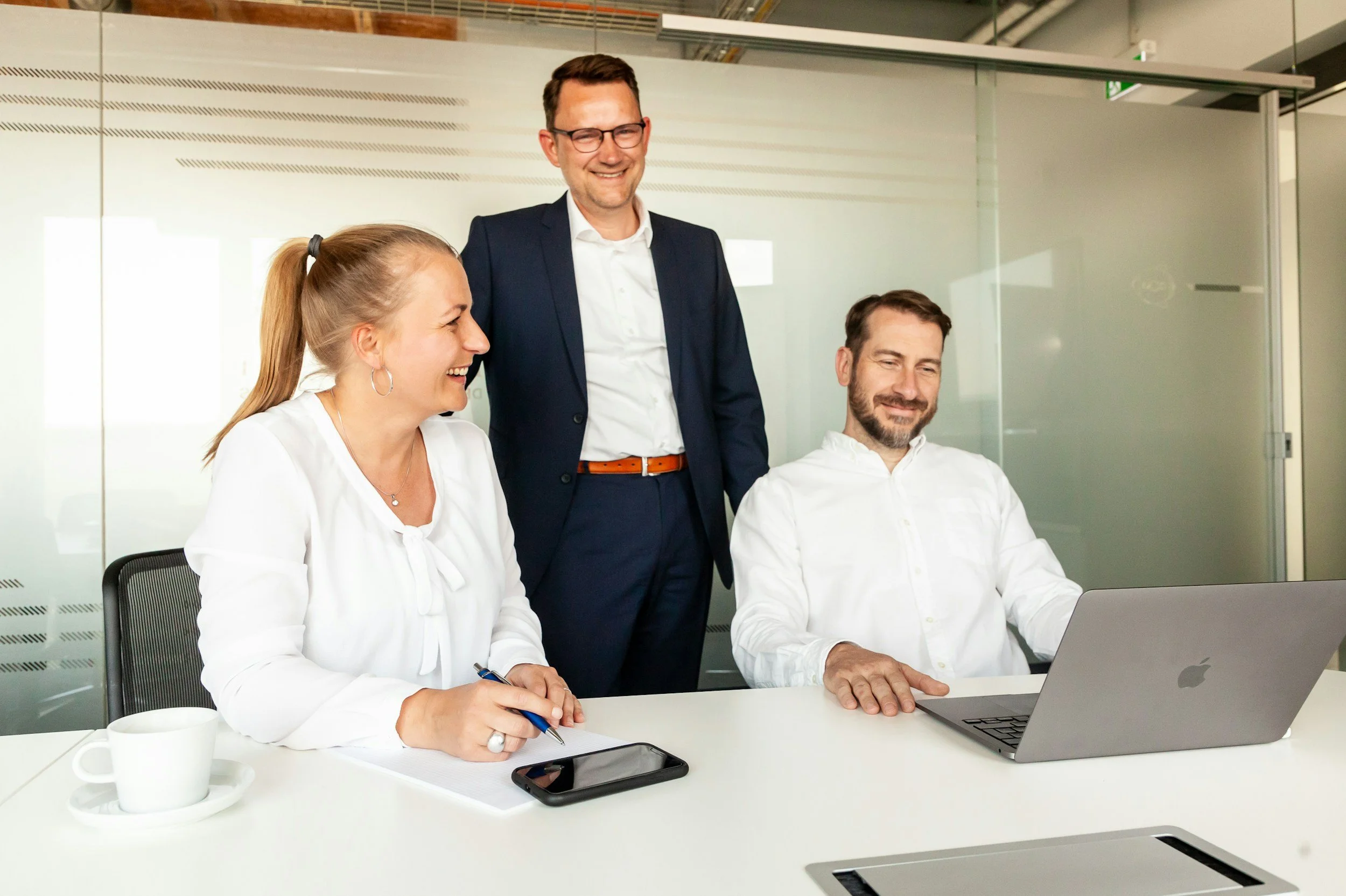 Four professionals in a modern office at a meeting. Two women and two men, one standing and three seated. They are smiling and working on a laptop and notes.