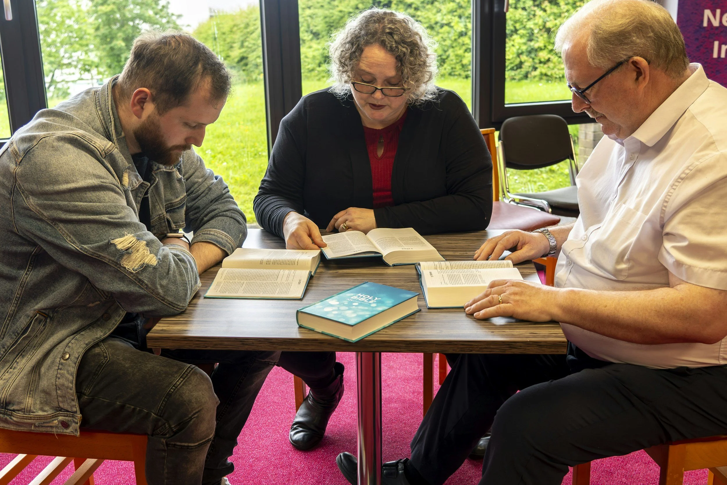 Three people sitting at a table reading and studying the Bible together in a bright room with large windows showing green outdoor scenery.