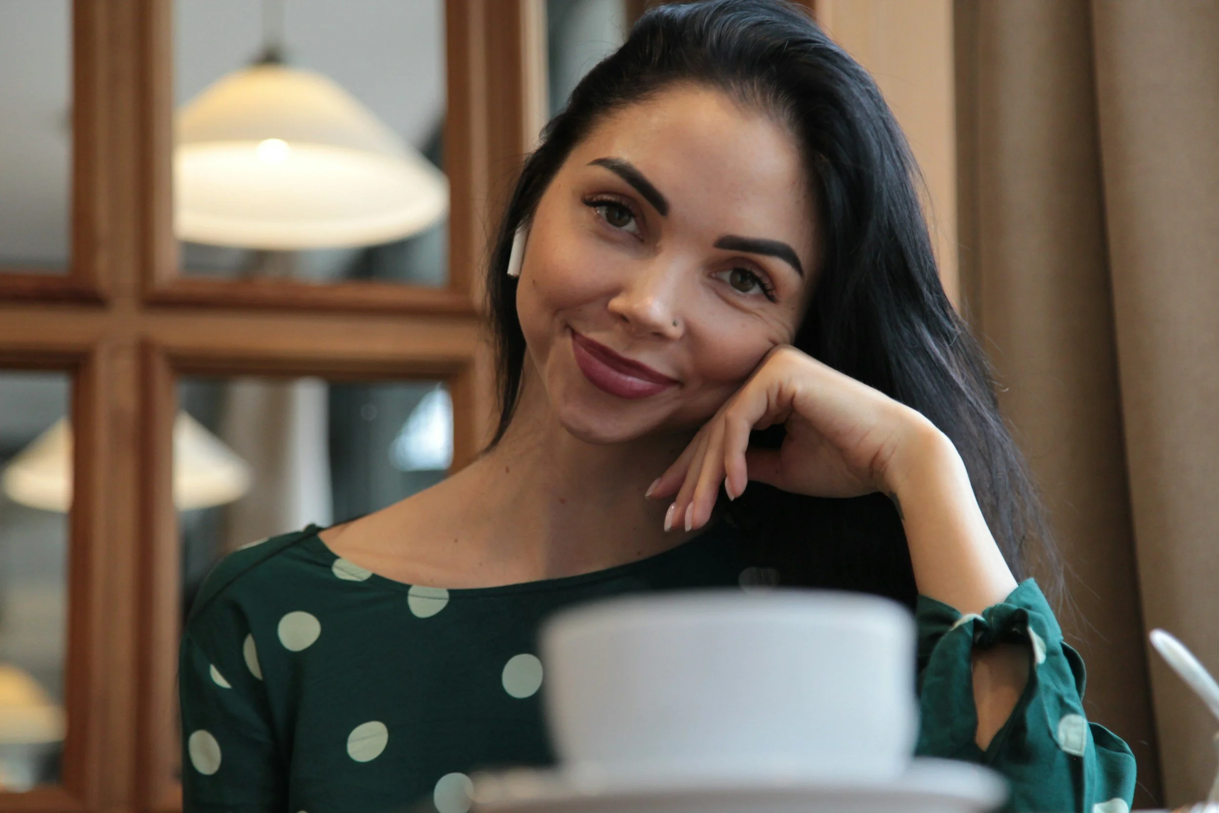 A woman with long dark hair, wearing a green polka dot shirt and AirPods, sitting at a table with a white mug and a pen, resting her chin on her hand and smiling softly.