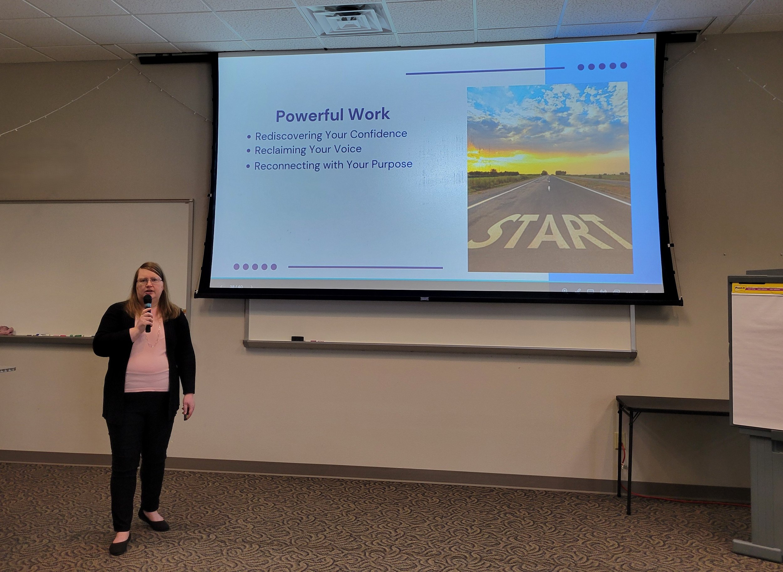 A woman holding a microphone standing in front of a large projection screen in a classroom or conference room. The slide on the screen reads 'Powerful Work' and lists three points: Rediscovering Your Confidence, Reclaiming Your Voice, Reconnecting with Your Purpose. There is a photograph of a road with a sunset or sunrise in the sky.