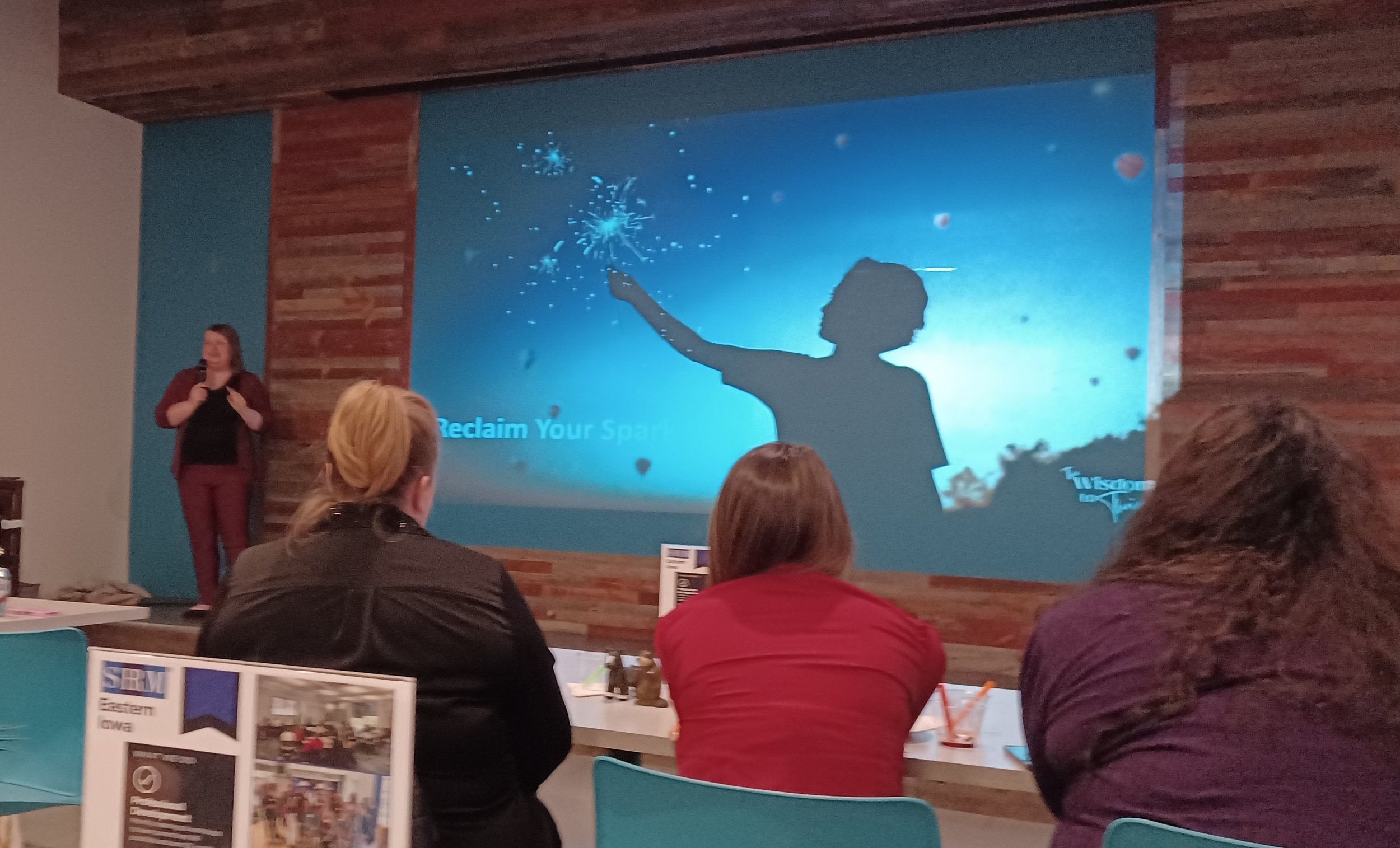 Michelle Pezleygiving a presentation in a conference room with a wooden wall. The slide on the screen shows a silhouette of a woman holding a sparkler against a blue sky. There are audience members seated, some with brochures or flyers on the tables.