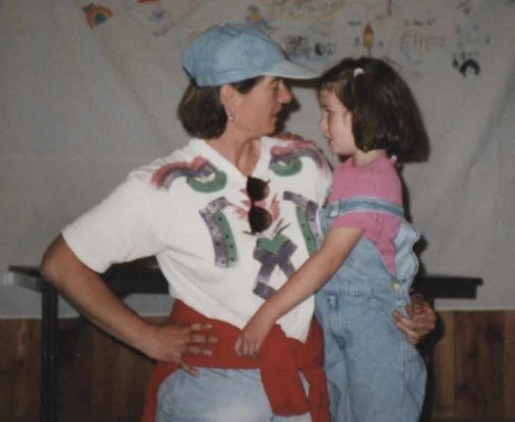 A woman in a colorful shirt and a blue cap holding a young girl in a pink shirt and overalls, engaging in a close, affectionate moment indoors with child's artwork on the wall in the background.