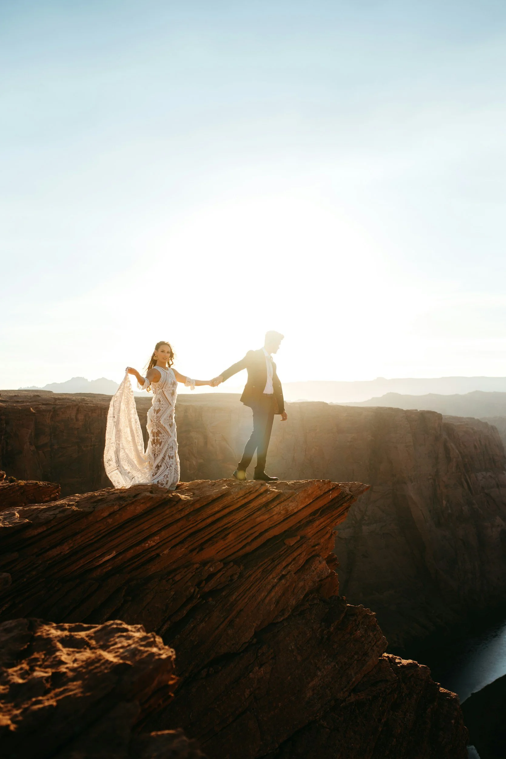 A couple holding hands on the edge of a cliff during sunset, with expansive canyon landscape in the background.