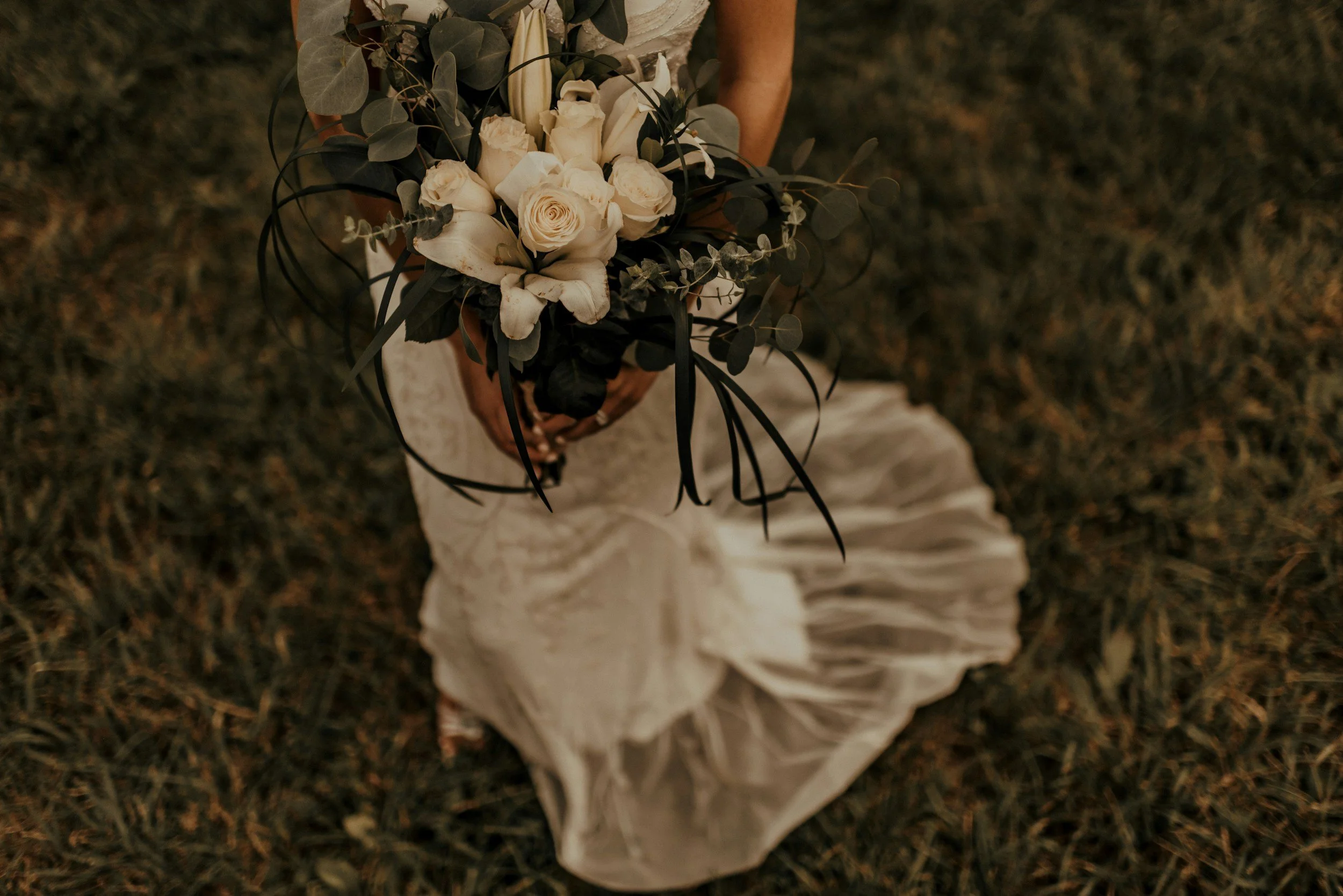 A person in a white dress holding a bouquet of white roses, greenery, and other flowers, standing on grass.