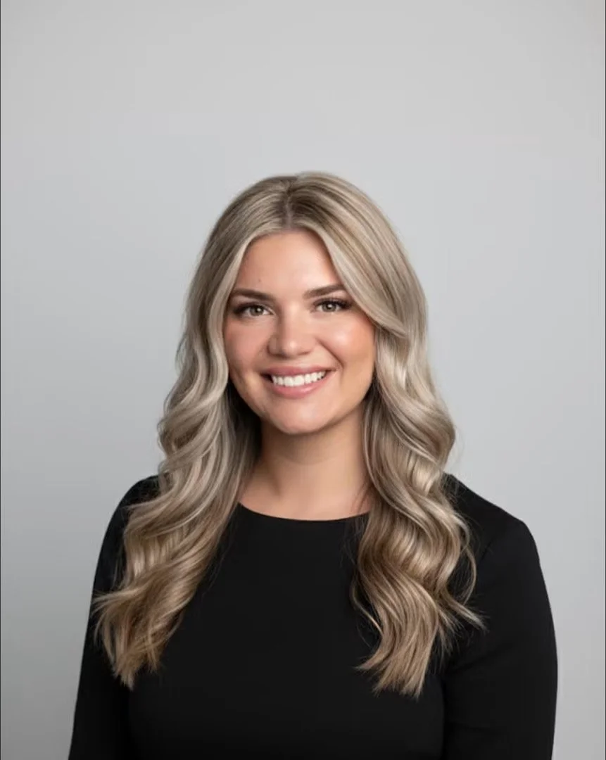 A woman with long, wavy blonde hair, smiling, wearing a black top, against a plain gray background.