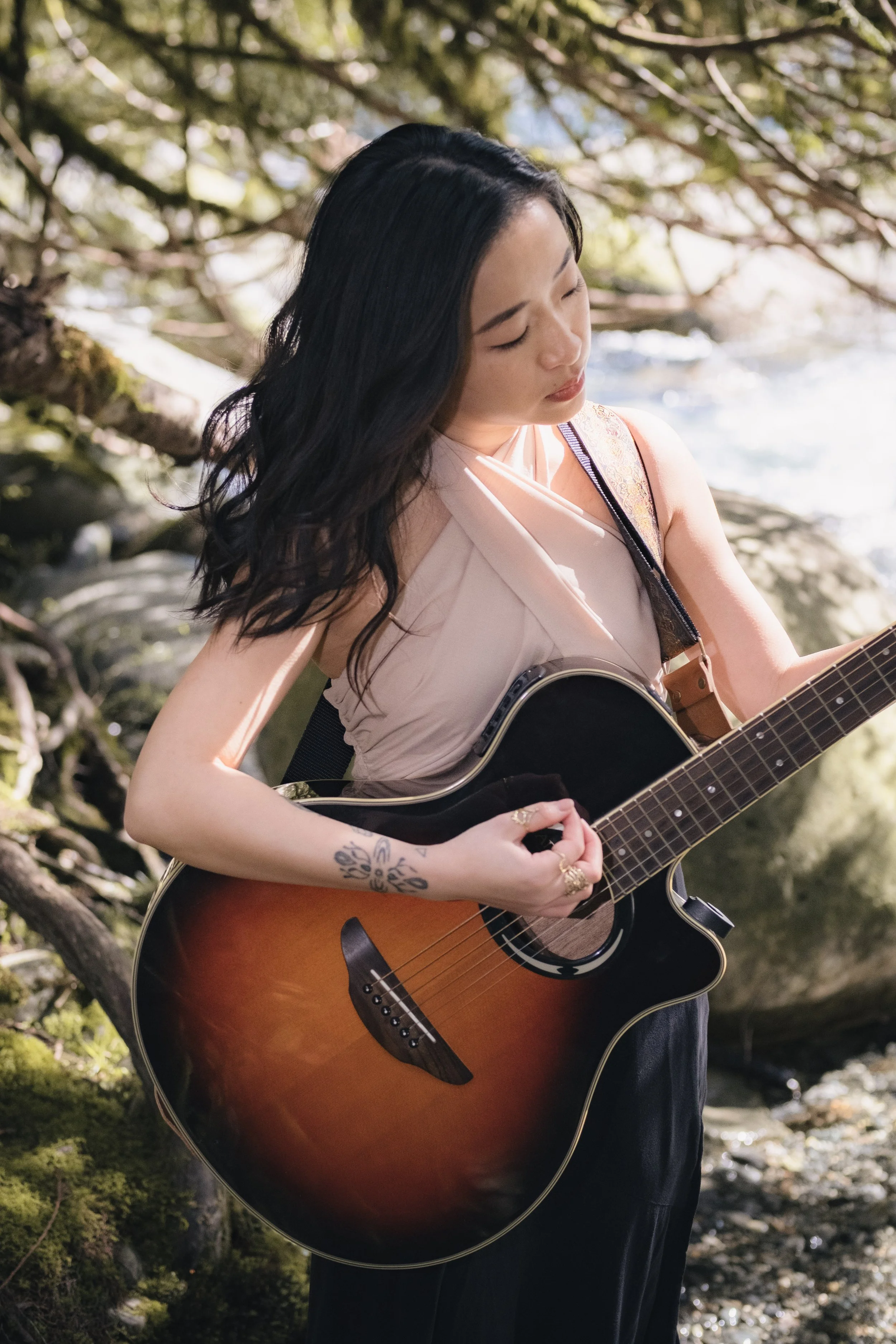 A woman with long dark hair, wearing a sleeveless top, playing an acoustic guitar outdoors in a natural setting with trees and sunlight.