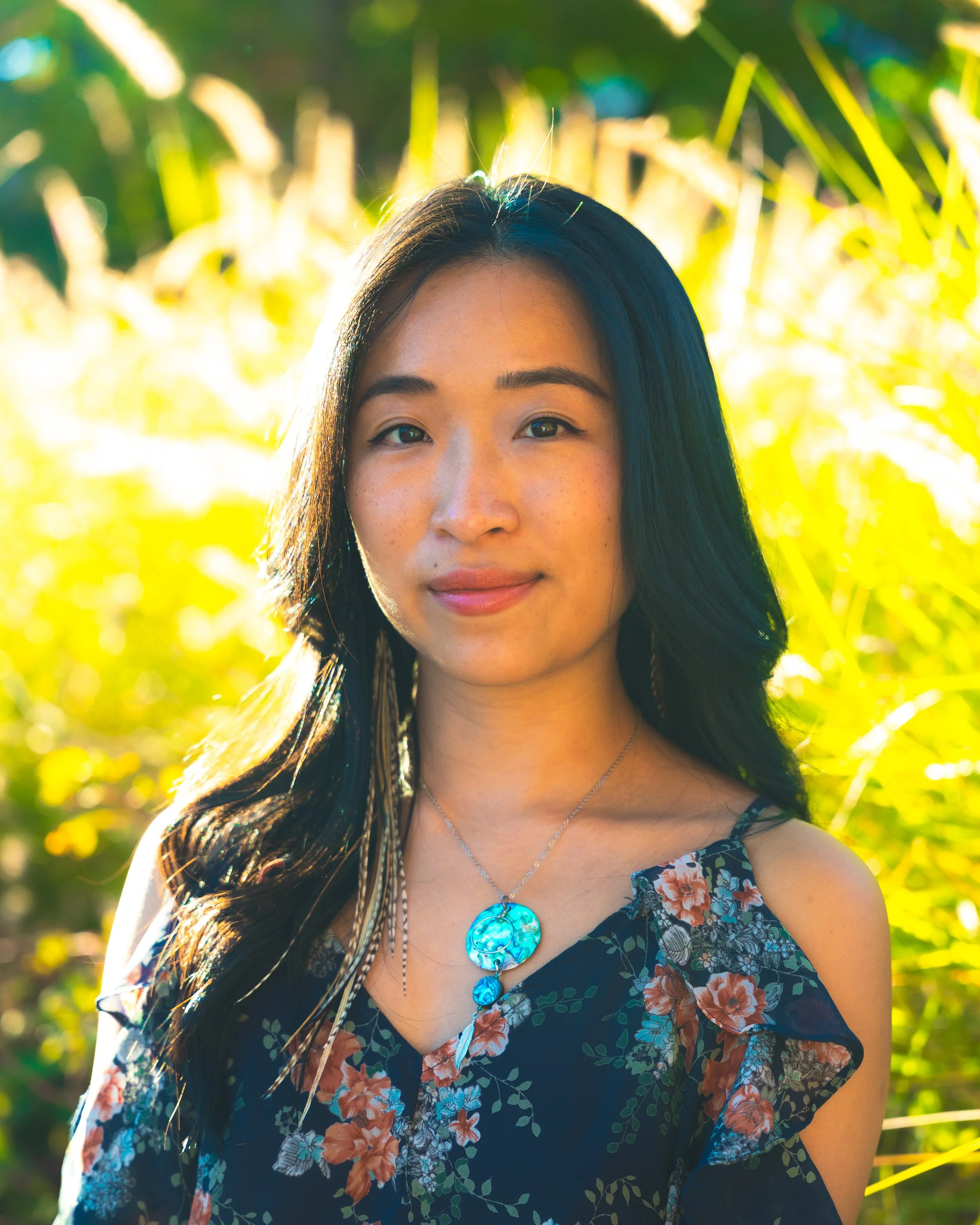A young woman with long dark hair standing outdoors in a garden or field of tall plants, wearing a floral dress and turquoise jewelry, with sunlight illuminating her face and hair.
