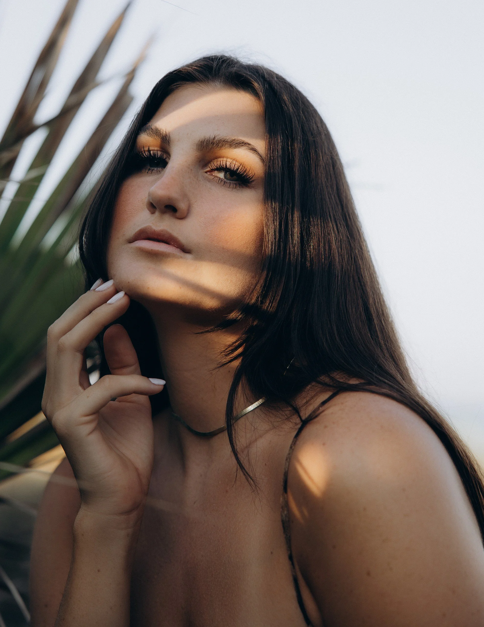Esthetician Kortni Gilson standing with hand on chin in front of a palm tree backdrop.