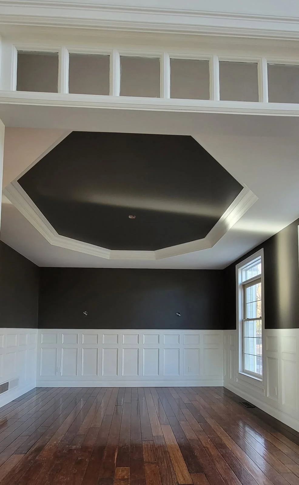 Professional painter in chester springs painted an Empty room with dark accent wall, white wainscoting, large window, and wood flooring, with a ceiling featuring a black octagonal recess.