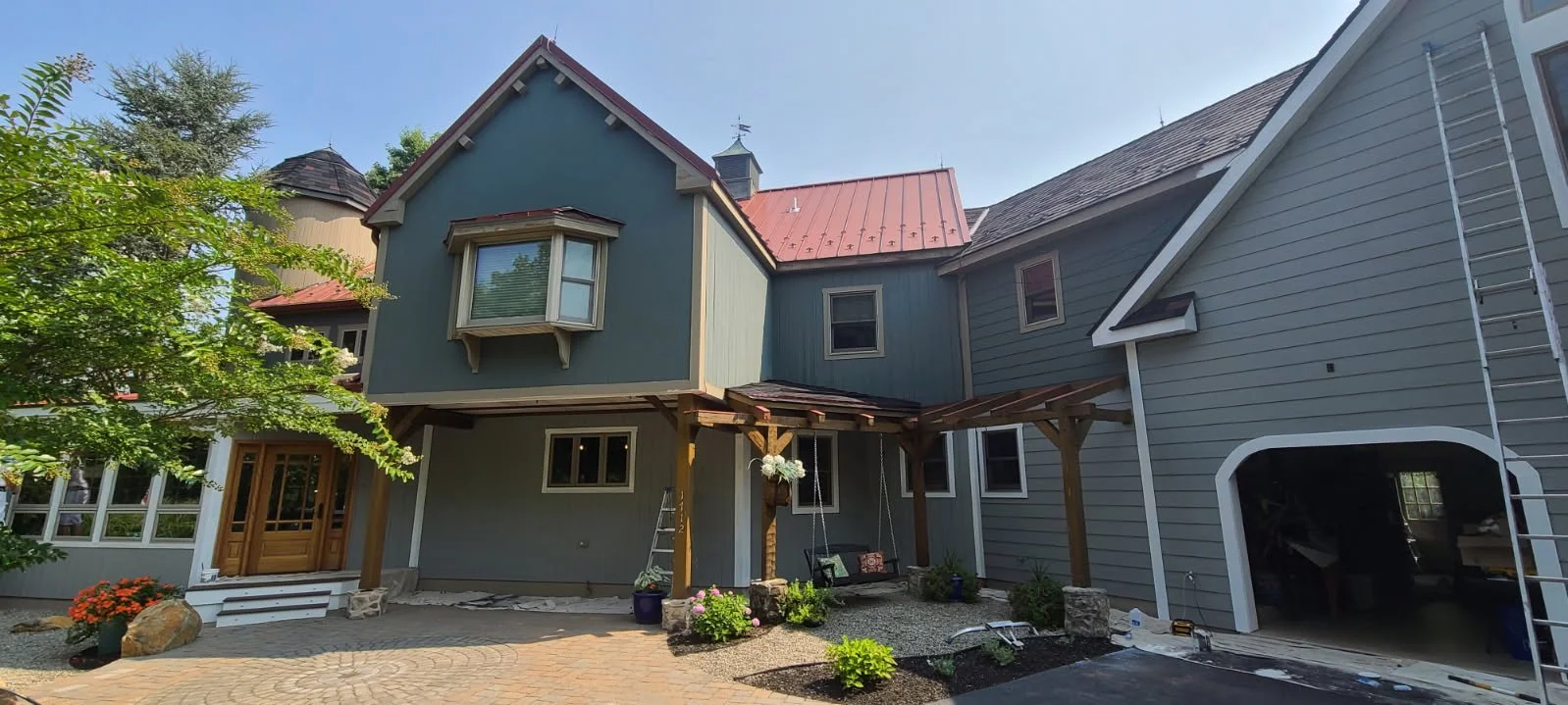 A multi-story house under construction with a wooden porch, swing, plants, and a ladder, featuring a mix of blue and gray siding and a red metal roof, with a tree in the foreground and a clear sky.