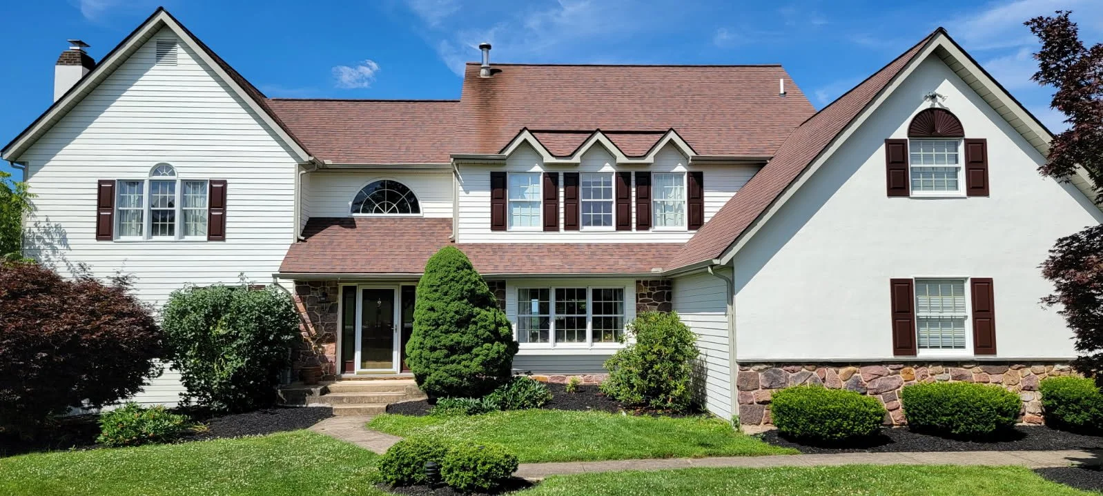 Professional painter in chester springs paints a large, two-story house with white siding, brown shutters, and a red-brown roof.