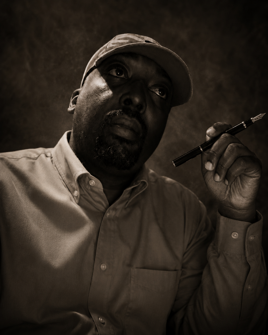 A black man wearing a baseball cap and dress shirt, holding a pen near his face, looking thoughtfully into the distance in a sepia-toned portrait.