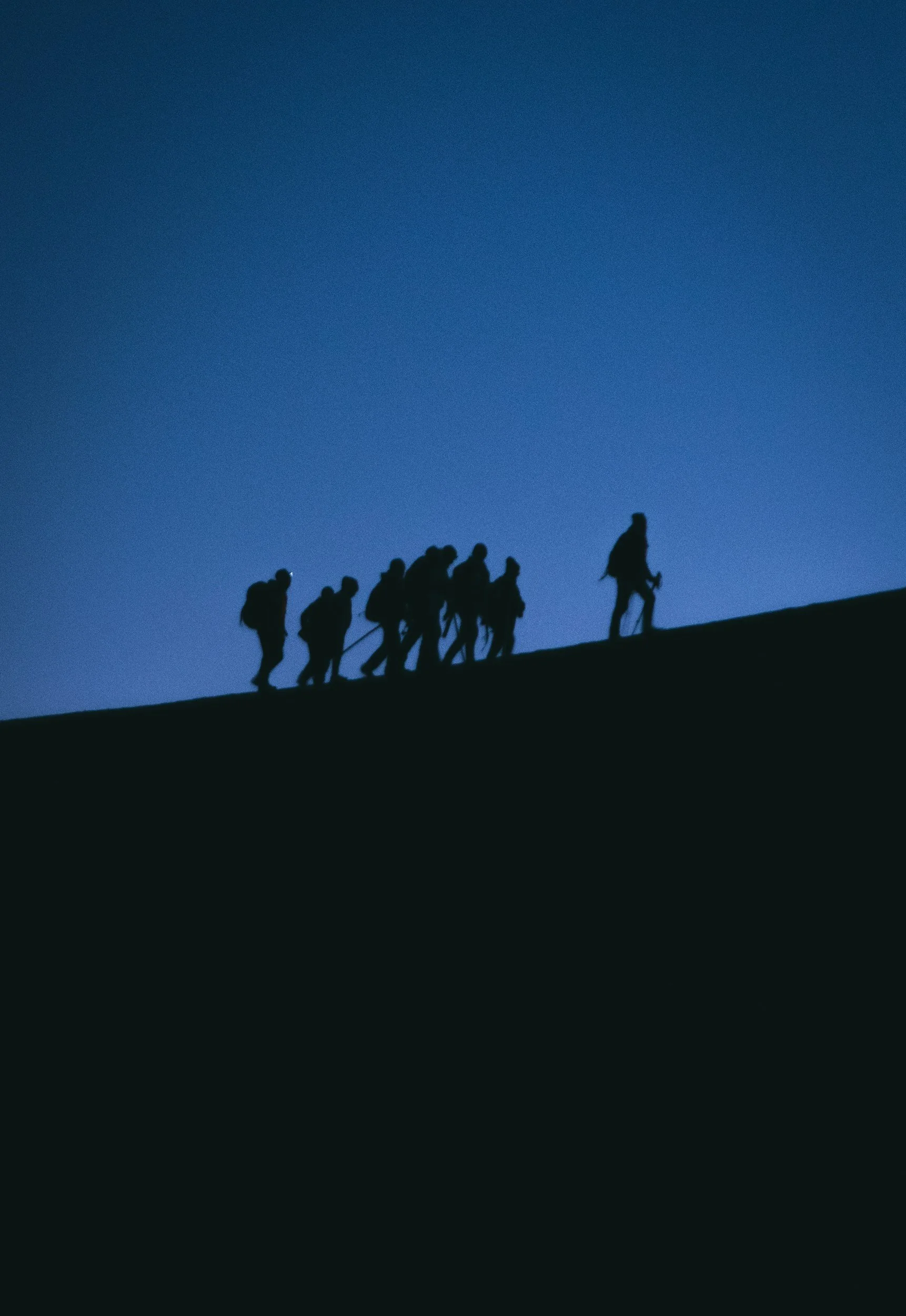 Groupe de personnes marchant en file sur un sommet de montagne au crépuscule ou à l'aube, silhouette noire contre un ciel bleu profond. Le groupe est mené par une personne en avant.