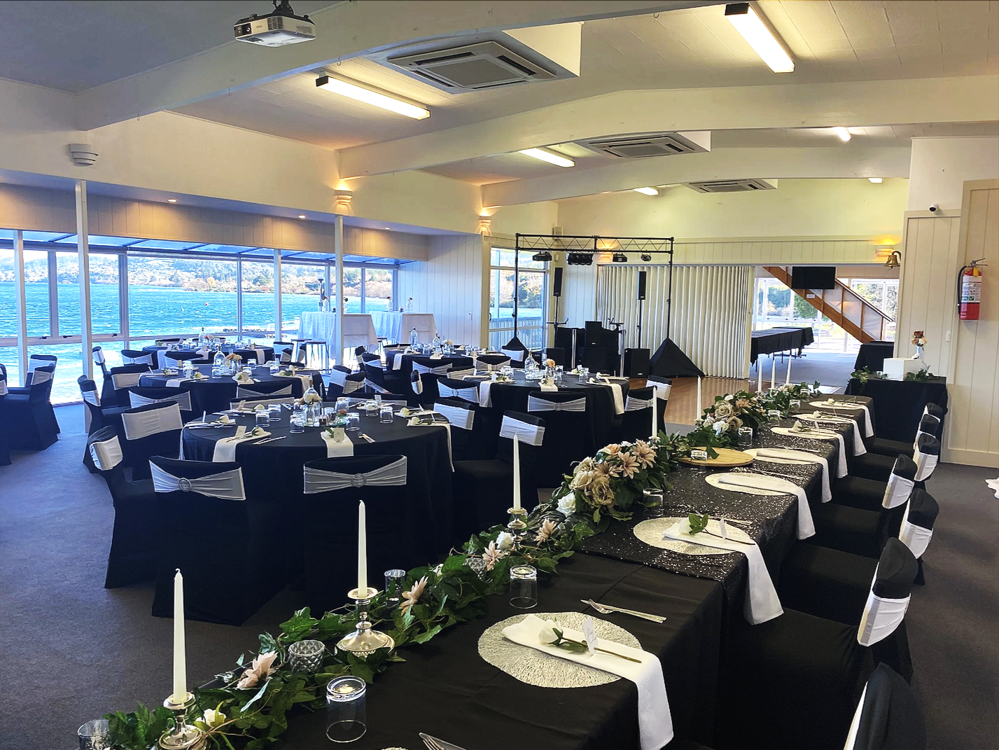 A decorated banquet in The Lake Taupo Yacht Club with tables set for an event, overlooking Lake Taupo  through large windows, with floral centerpieces and black and white table settings.