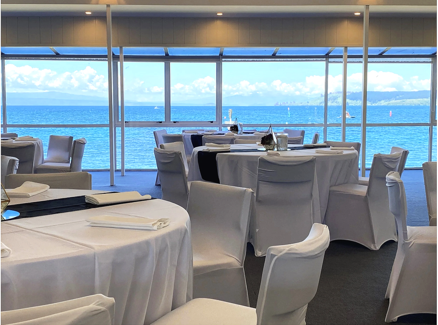 Elegant dining area In Lake Taupo Yacht Club with round tables covered in white tablecloths, set with napkins, overlooking a large window with a view of the lake  and sailboats.