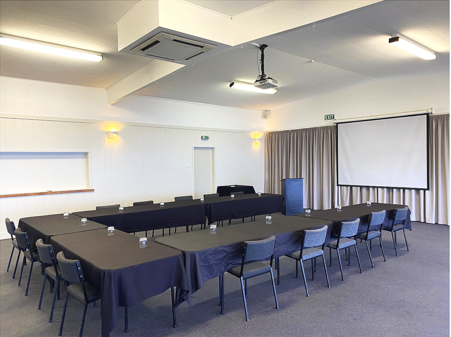 Empty conference room with U-shaped table arrangement, black tablecloths, blue chairs, a projector screen, and a podium, with gray curtains, white walls, and ceiling lights at Taupo yacht club.