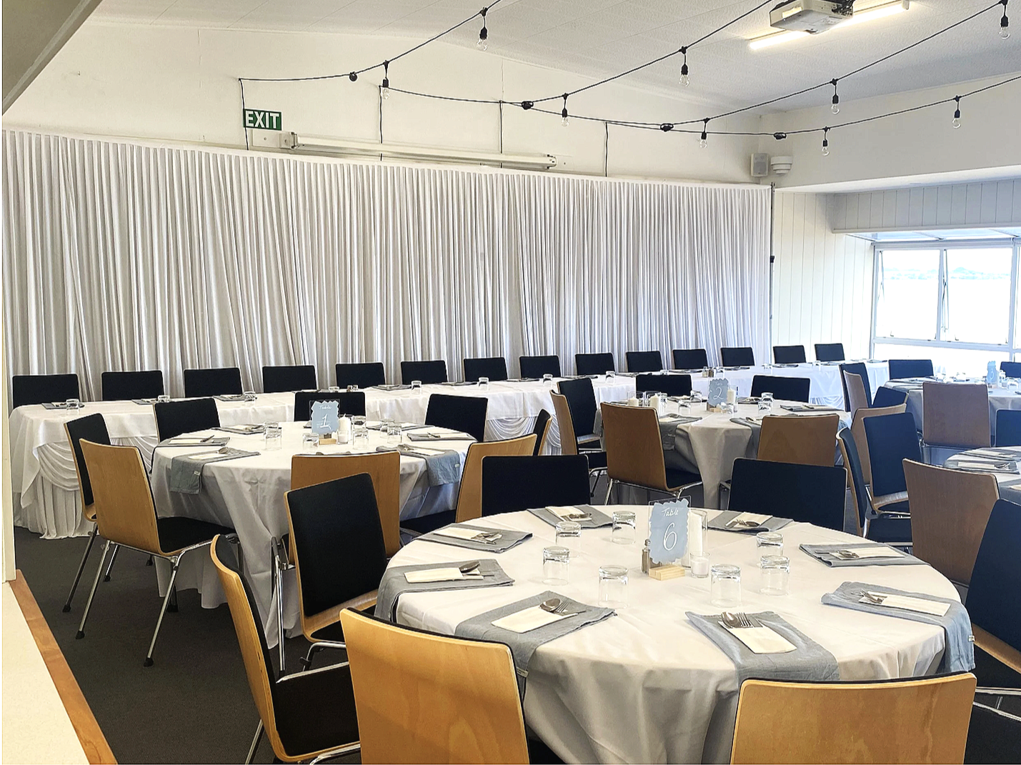 Wedding  set up with round tables covered in white tablecloths, each with napkins, utensils, and glasses. There is a white curtain on the wall, string lights hanging from the ceiling, and large window allowing natural light.