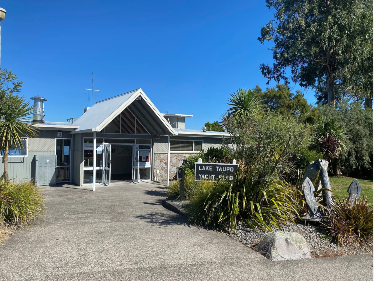 Entrance to Lake Taupo Yacht Club, with a sign, surrounded by plants and trees, under a clear blue sky.