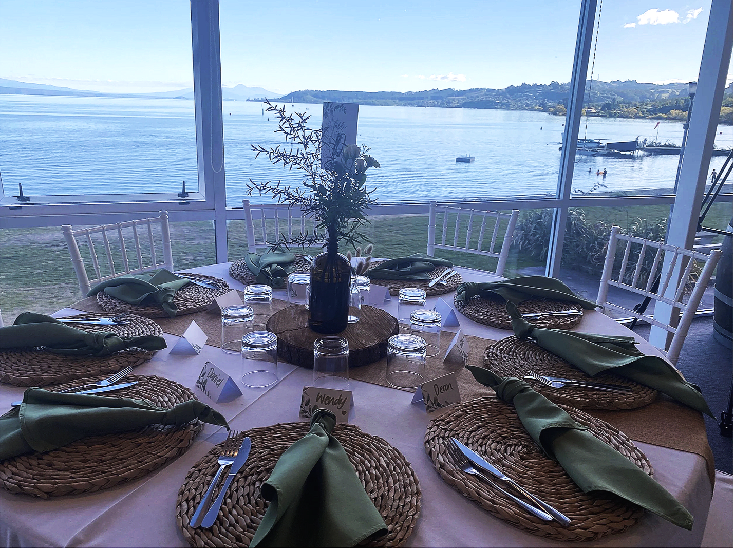 A round dining table set for eight, with woven placemats, green napkins, silverware, and upside-down glasses, positioned near large windows with a view of lake Taupo and distant land.