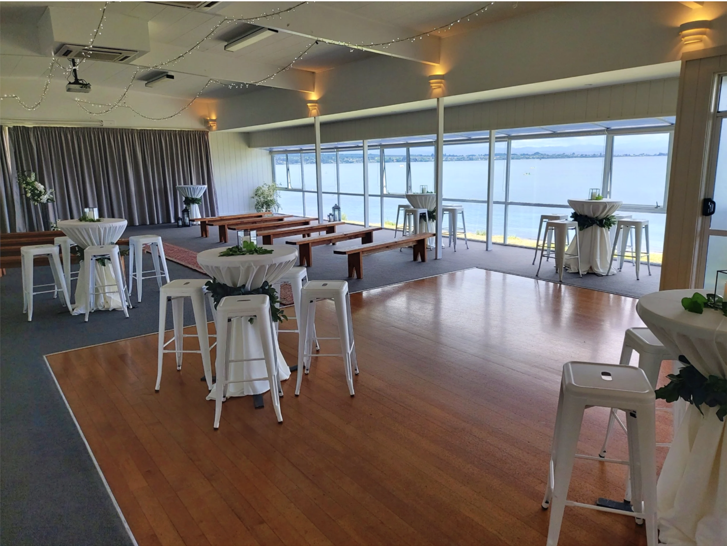 Event space decorated with high-top tables and benches overlooking a body of water through large windows, with string lights hanging from the ceiling.