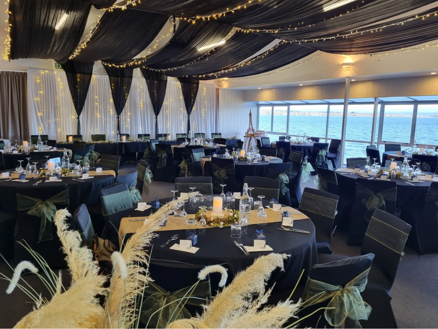 Elegant banquet hall decorated with black tablecloths, tied with green bows, and a central candle centerpiece. The hall features large windows, navy blue ceiling drapery with string lights, and white curtains with additional string lights. Tables are