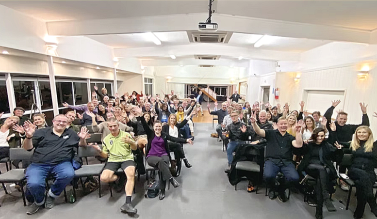 At Lake Taupo Yacht Club a large group of people in a spacious, well-lit room, sitting on chairs arranged in rows, smiling and waving at the camera during a social or corporate event.