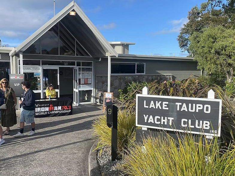 Entrance of Lake Taupo Yacht Club with a sign, surrounded by plants, with people standing outside on a sunny day.