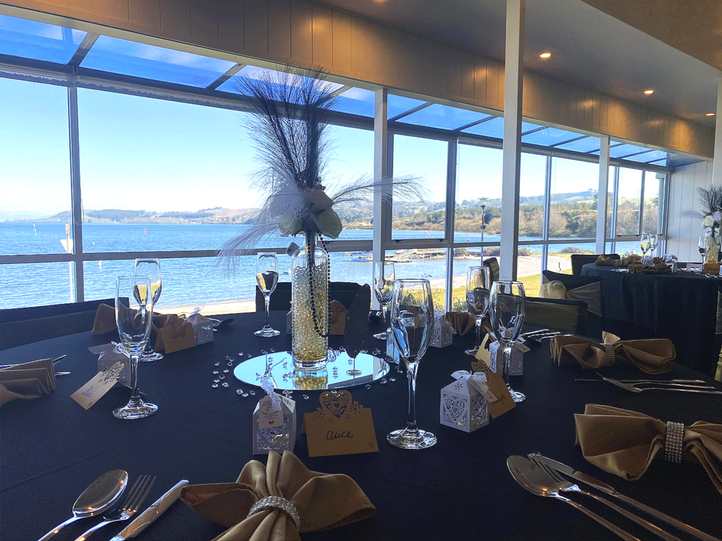 Elegant table with black tablecloth, crystal glasses, silverware, and gold-colored napkins with rhinestone bands. Glass vase with white flowers and decorative beads, surrounded by small gift boxes and rhinestones. Large window overlooking Lake Taupo.