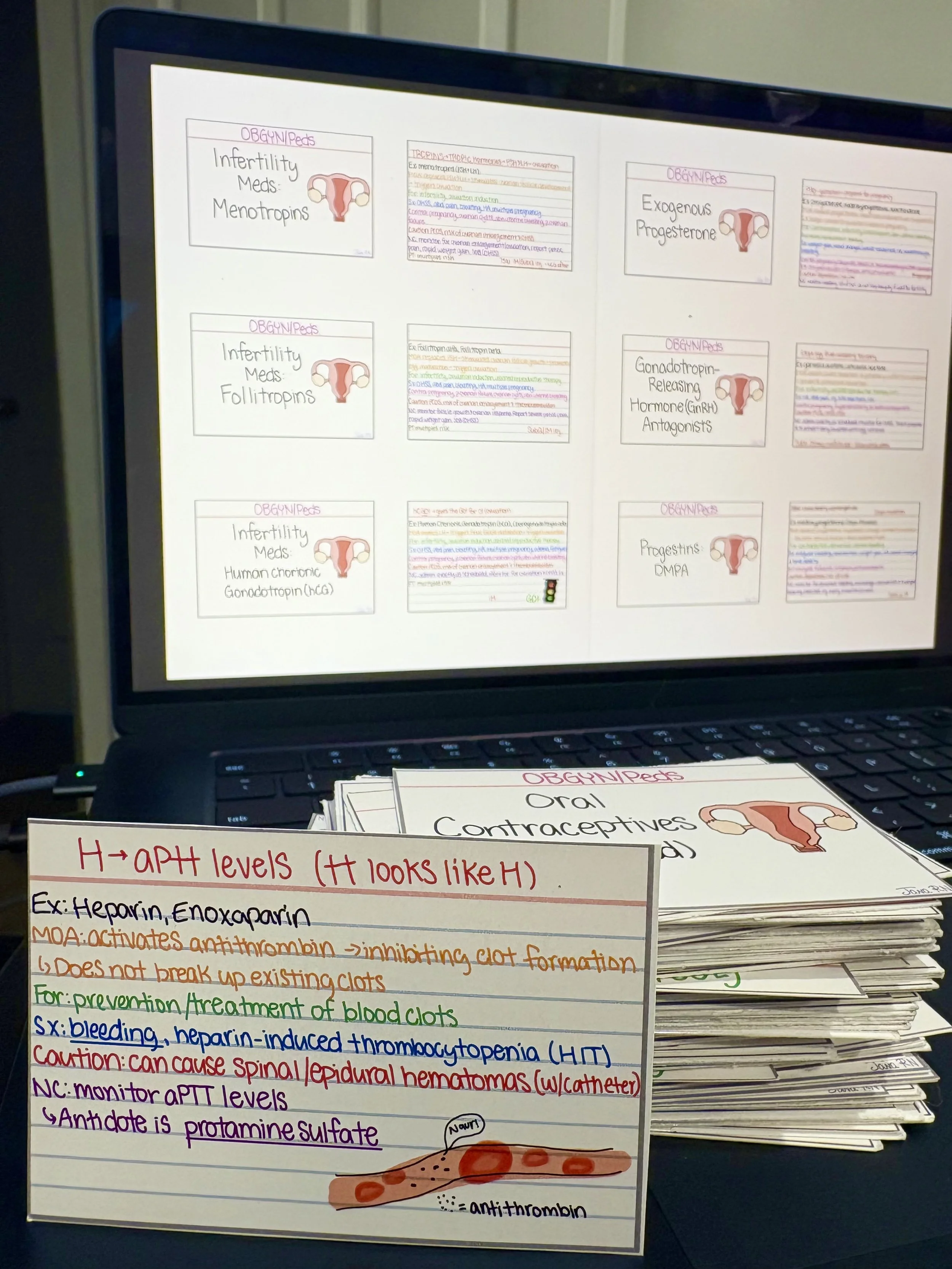 A computer monitor displaying typed notes about reproductive health, with additional handwritten notes and educational materials on blood clotting and anticoagulants placed in front of the screen.