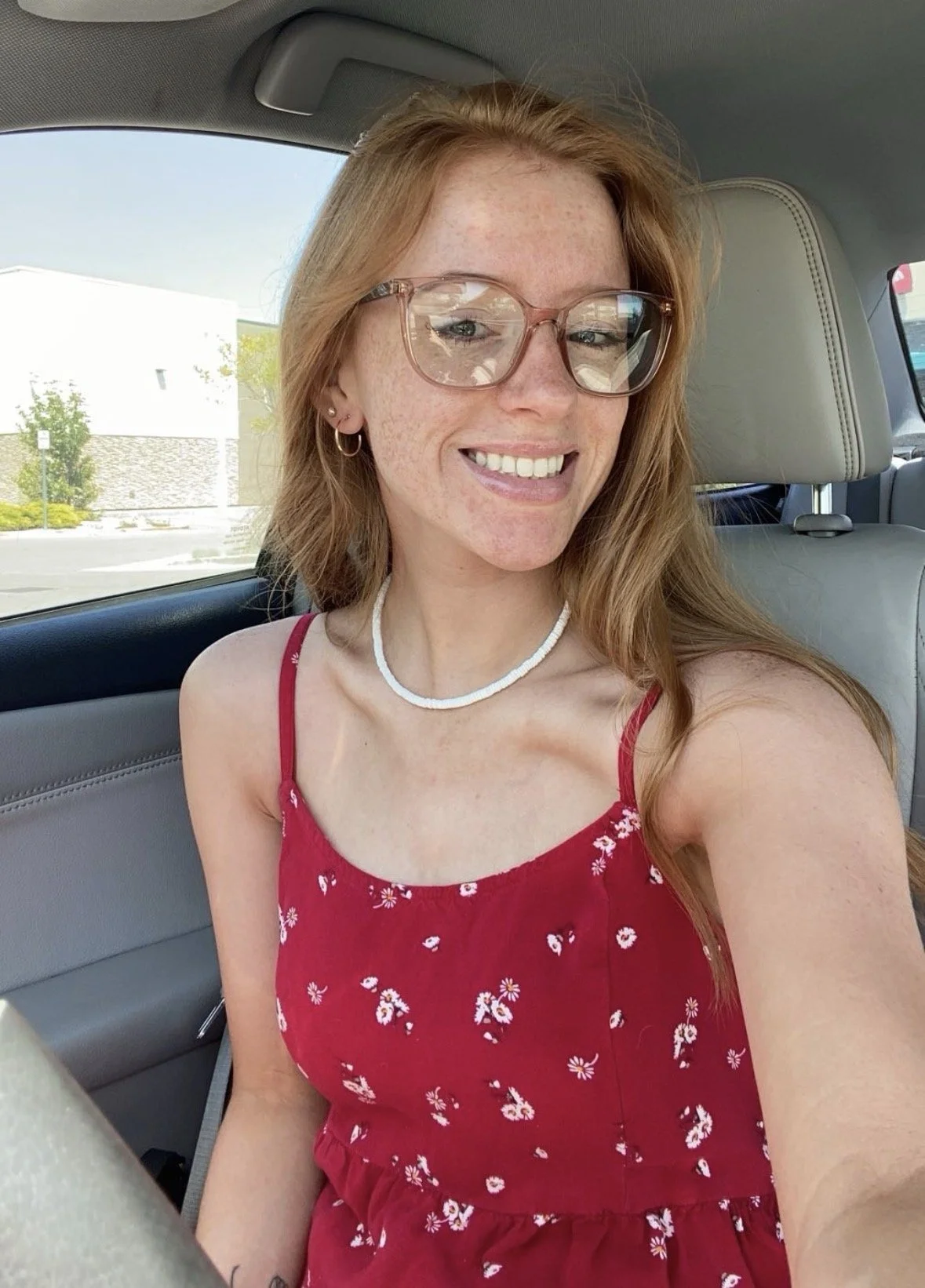 A young woman with long red hair and glasses sitting in the back seat of a car, smiling at the camera. She is wearing a red dress with white floral patterns, a white beaded necklace, and small earrings. The background shows a sunny day outside with a building and trees visible through the car window.