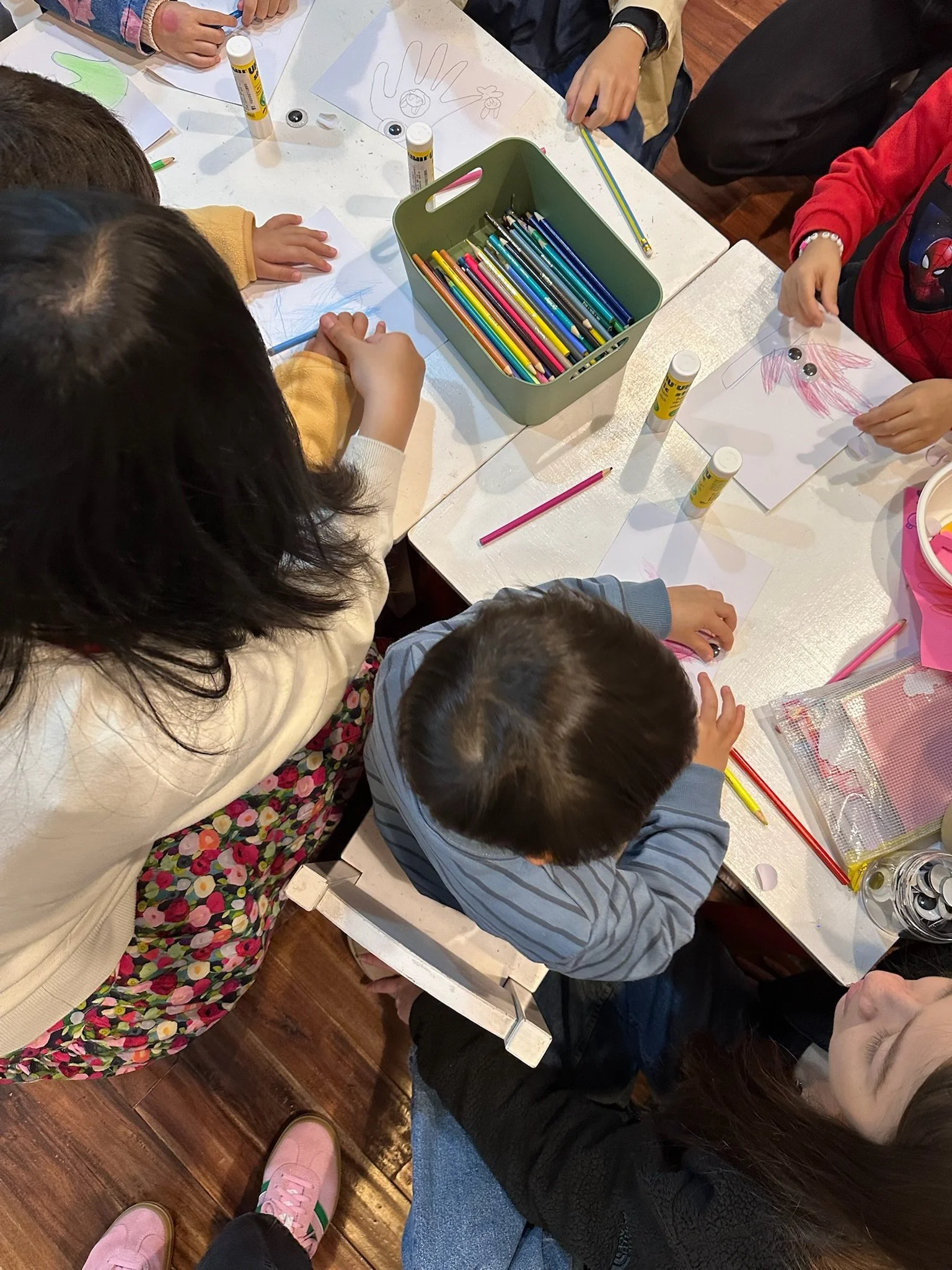 Children sitting around a table engaging in arts and crafts, drawing and coloring on paper, with colored pencils, glue sticks, and other supplies scattered on the table.
