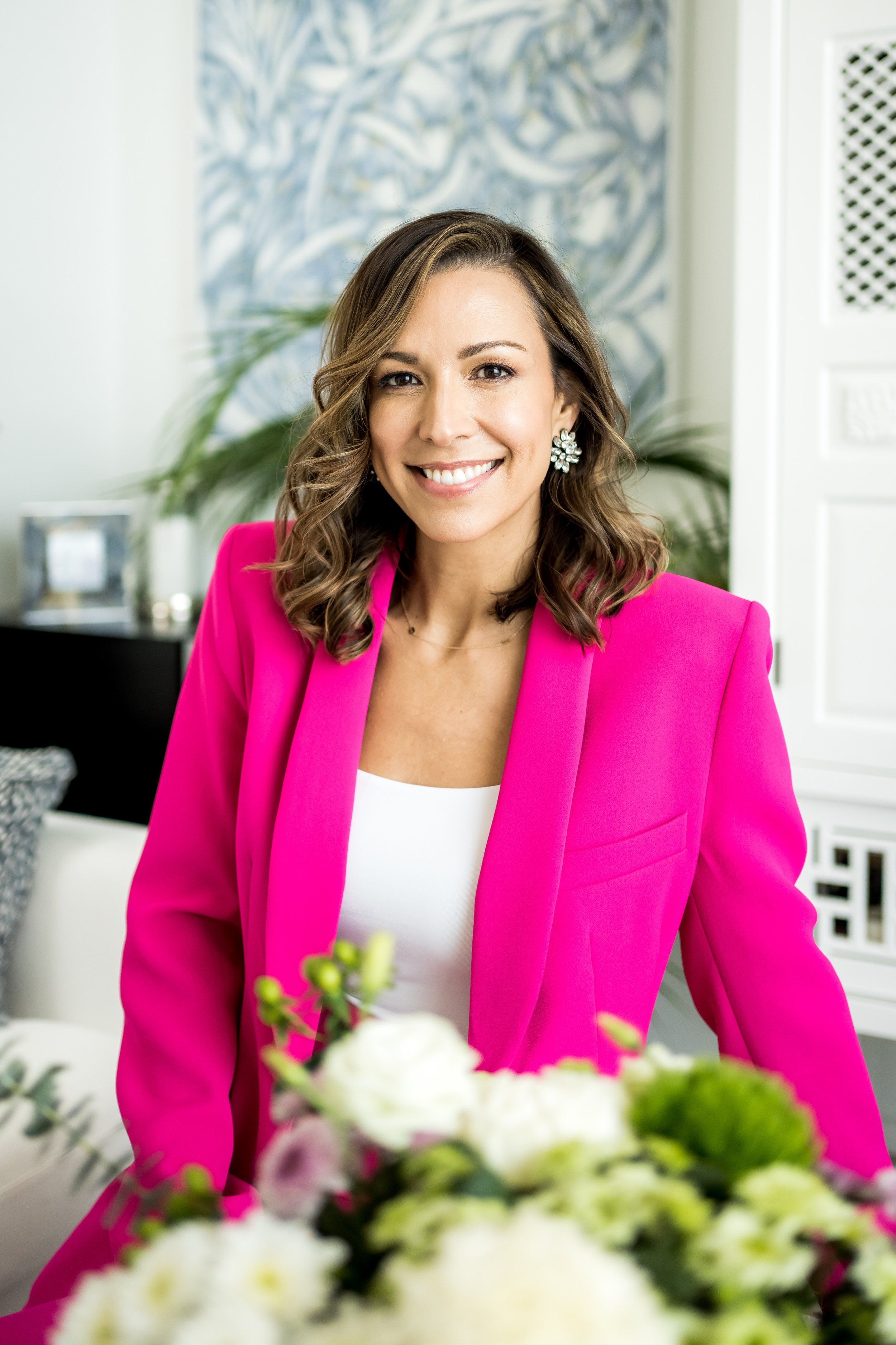 A woman with wavy brown hair and earrings, smiling, wearing a bright pink blazer and white top, sitting in a brightly lit room with artwork on the wall and plants in the background.