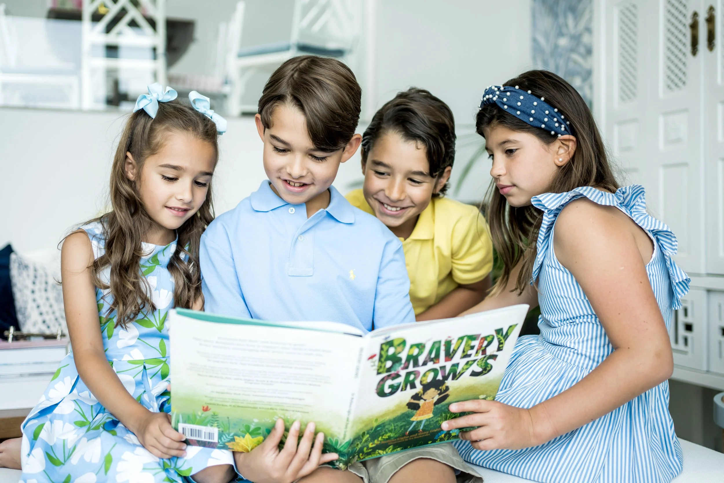 Four young children sitting together inside, looking at a colorful book titled "Bravery Grows." The children are smiling and appear engaged, with two girls wearing blue dresses and bows, and two boys in light-colored shirts. The background features a