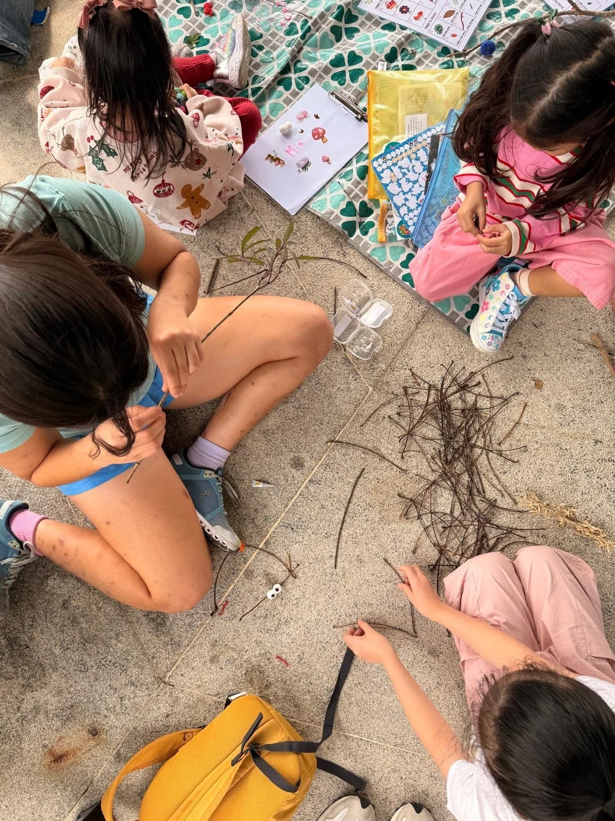 Children sitting on the ground, collecting and examining sticks and branches, with school supplies and a drawing on paper nearby.