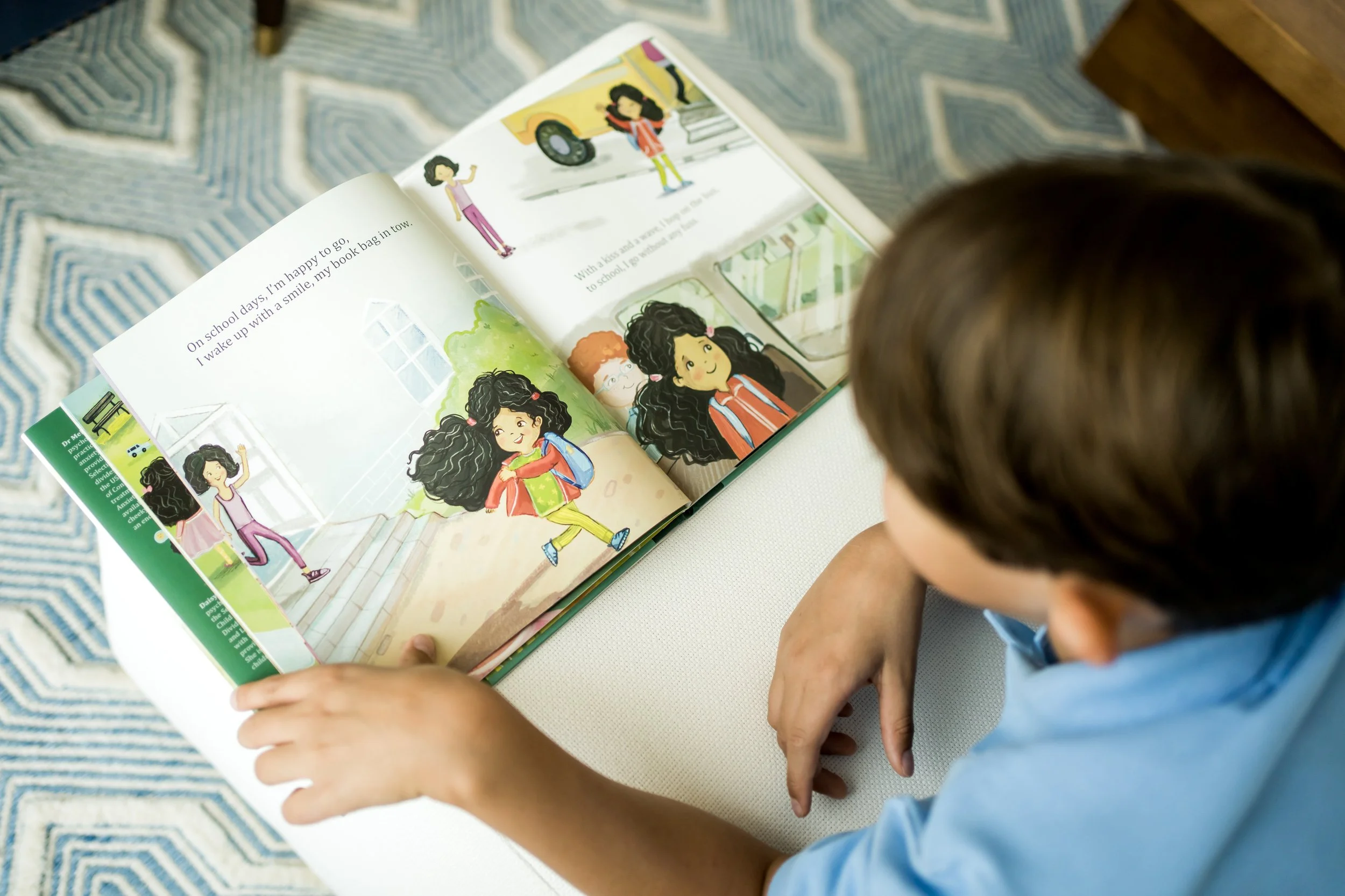 A child with curly hair wearing a blue shirt is looking at a colorful illustrated children's book on a white surface, with a blue and white geometric patterned rug underneath.