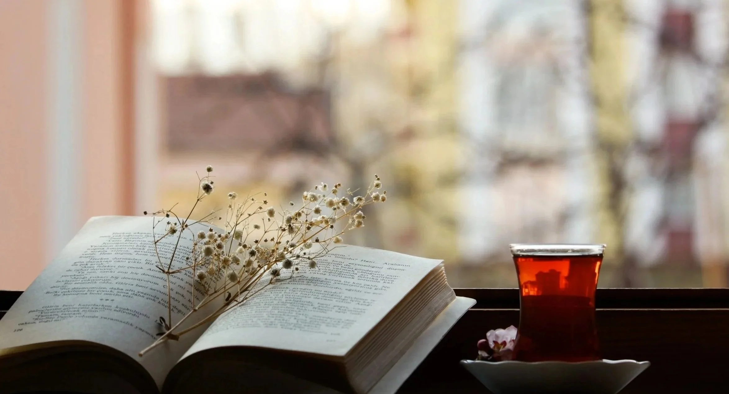 Open book with dried flowers on pages, a glass of tea, and a blurred window view in the background.