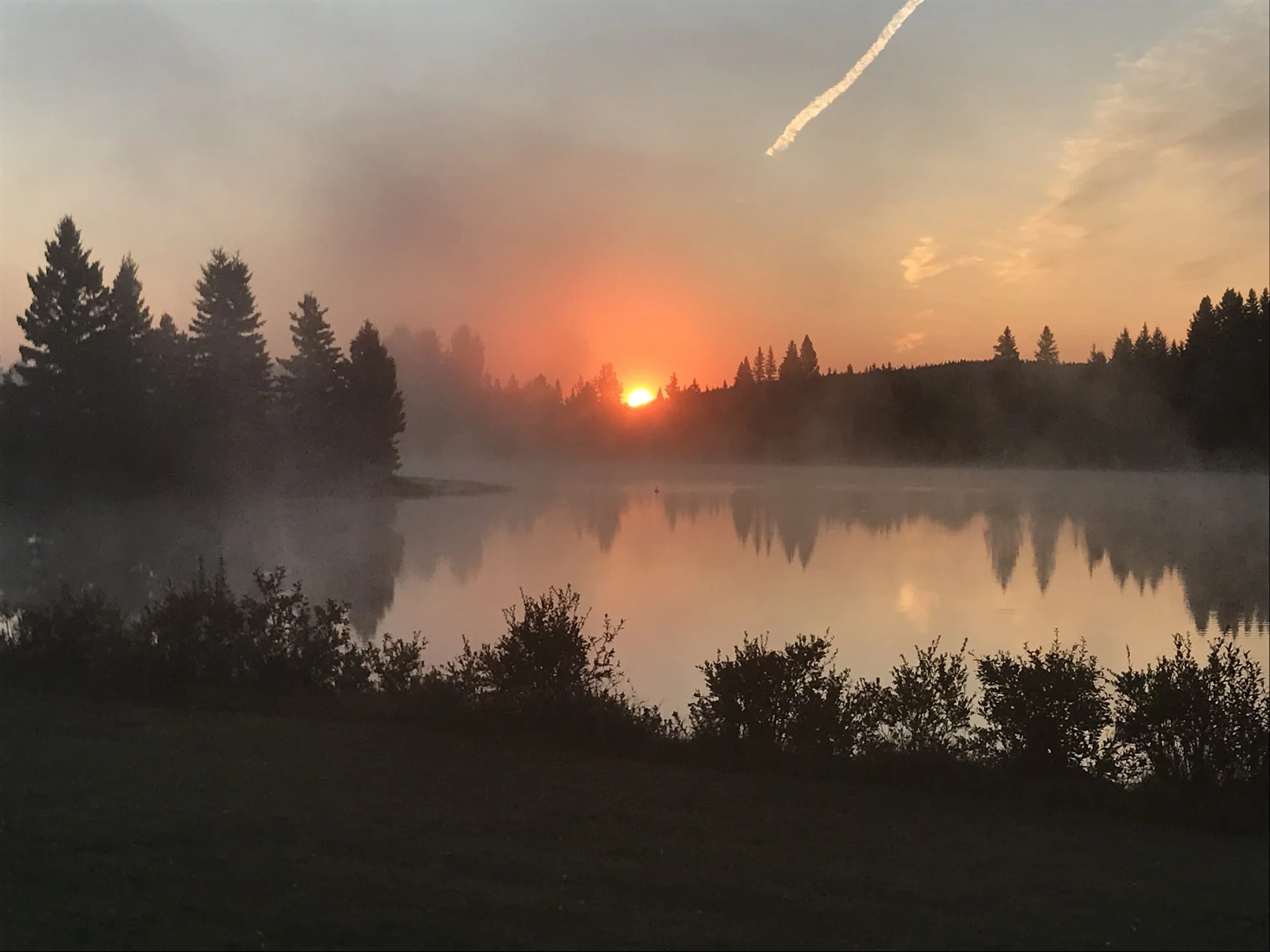 Sunset over a calm lake with trees along the shore, reflecting in the water, with a trail of smoke in the sky.