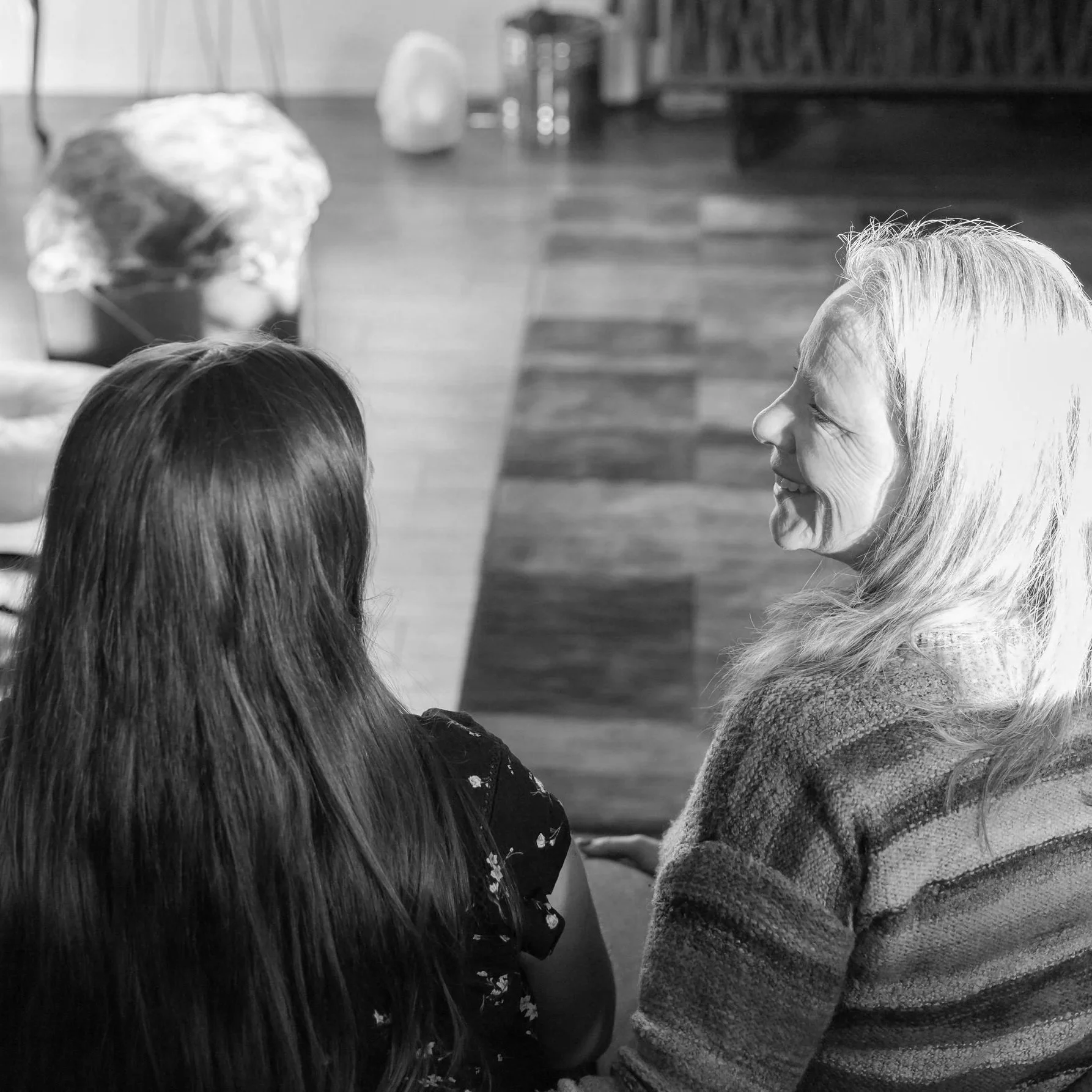 A woman with long hair and a child facing away from the camera, sitting together and smiling, in a room with wooden flooring, bookshelves, and a blurred black-and-white background.