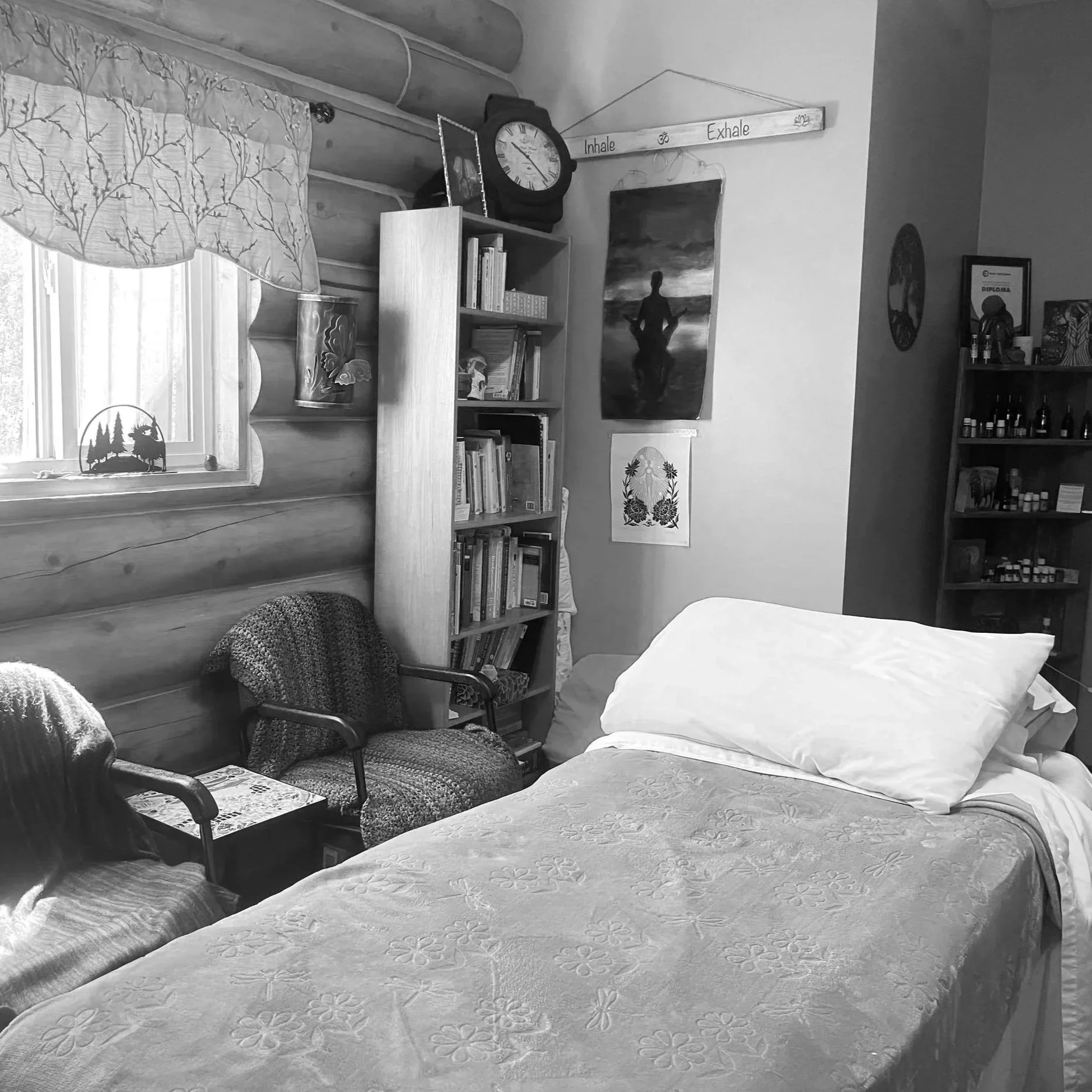 A bedroom with a bed covered in a floral patterned bedspread, and a wooden chair with padding and a newspaper on it. There is a window with a shade that has a tree silhouette decoration, and a bookshelf with items and books. On the wall, there is a framed diploma and a bookshelf with various bottles. A poster of a woman is hanging on the wall, and a clock with a black frame is on a shelf, along with a decorative plaque with the words 'Inhale' and 'Exhale' and symbols on a hanging wooden plaque.