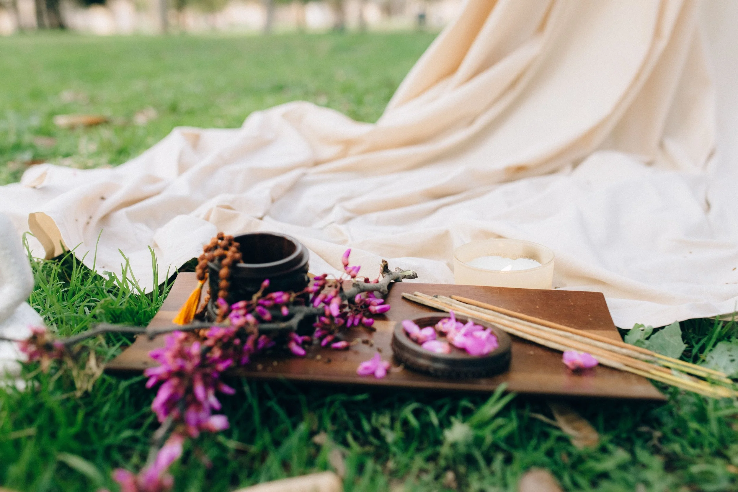 Close-up of a tabletop with pink flower petals, incense sticks, and small containers, set on grass with a white cloth and blurred background.