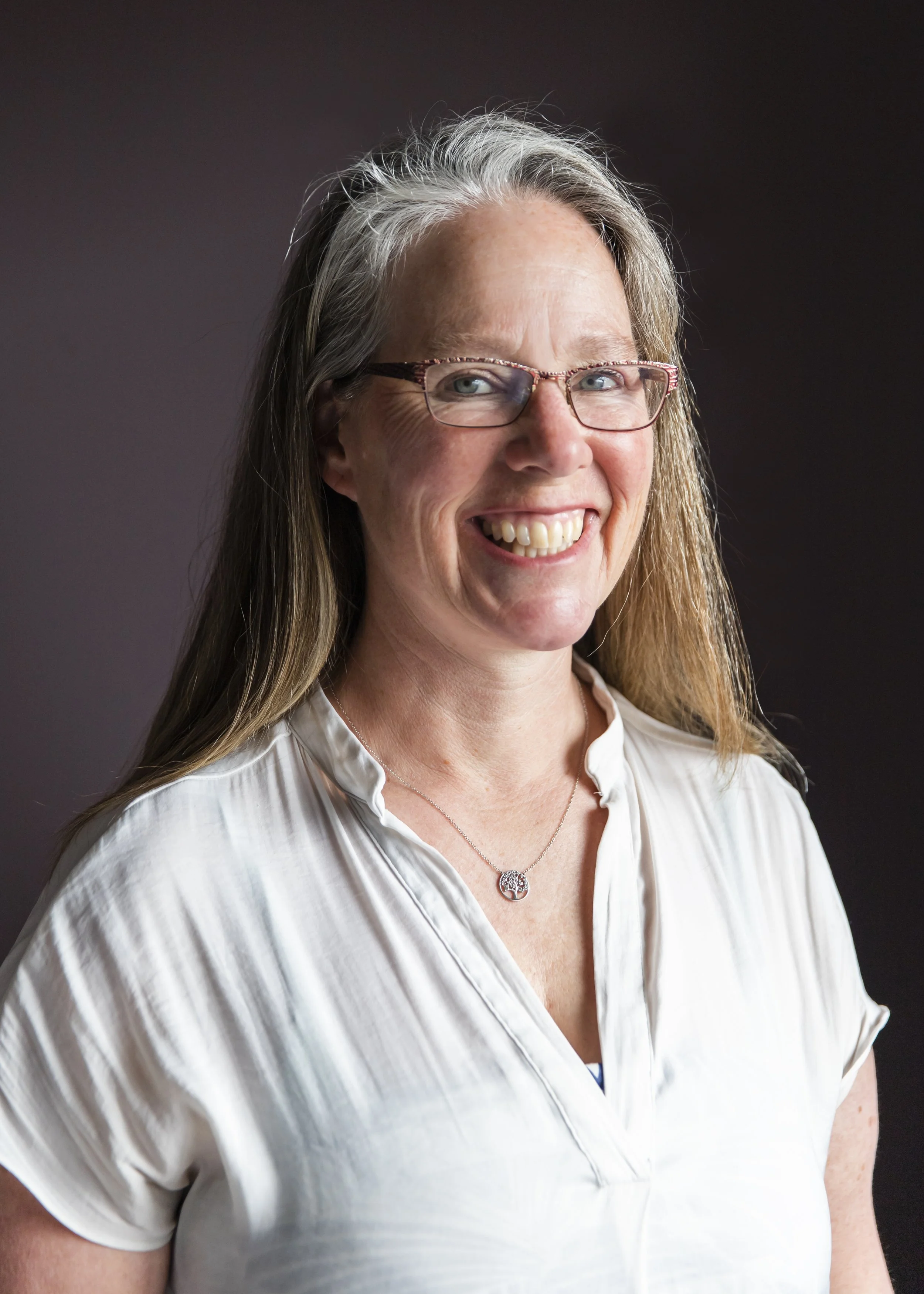 A smiling woman with glasses and long gray hair, wearing a white blouse and a necklace with a tree pendant, against a dark background.