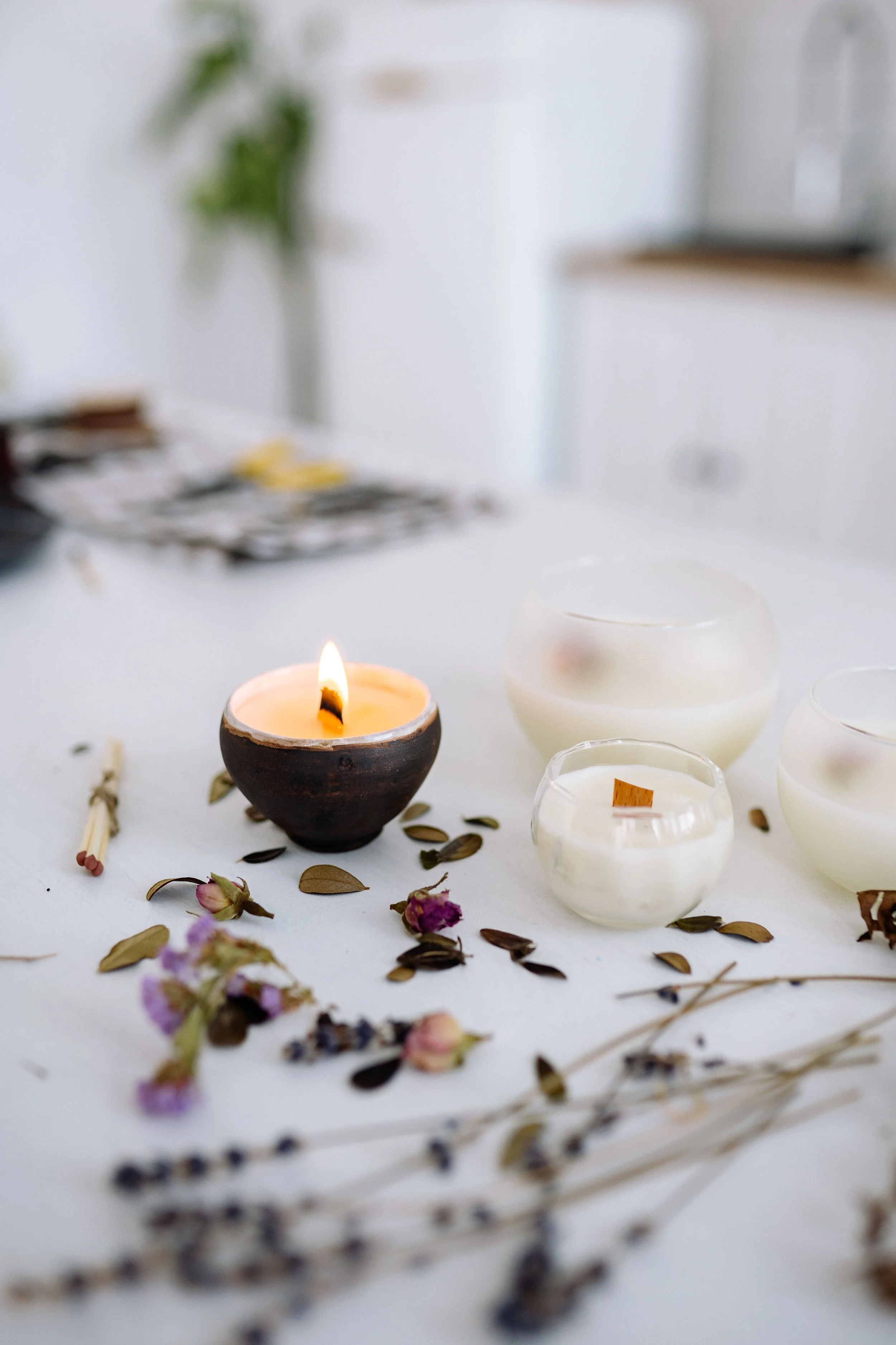 A partially burned candle in a small dark bowl, surrounded by empty glass candle holders, dried leaves, small flowers, and matchsticks on a white surface. In the background, there are blurred objects and a potted plant.