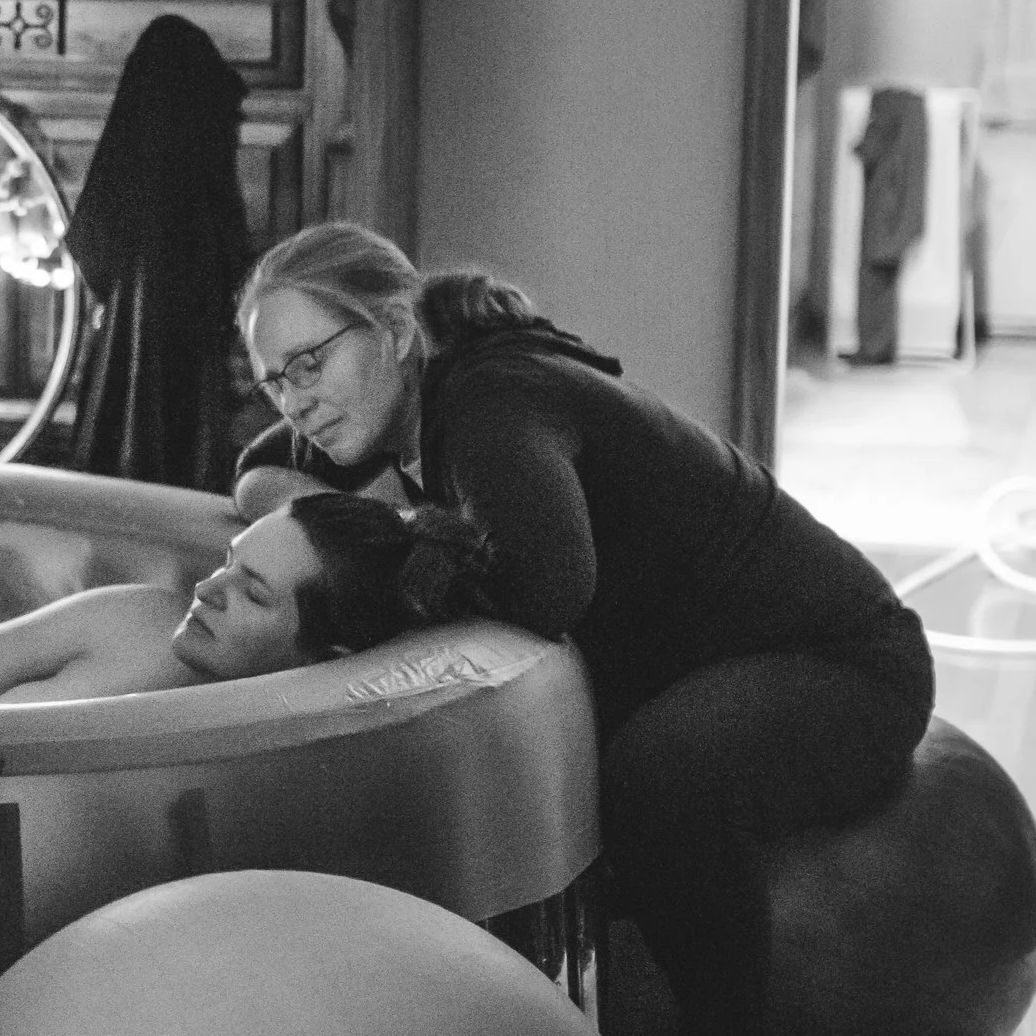 A woman with glasses leaning over a woman lying in a bathtub, both smiling.