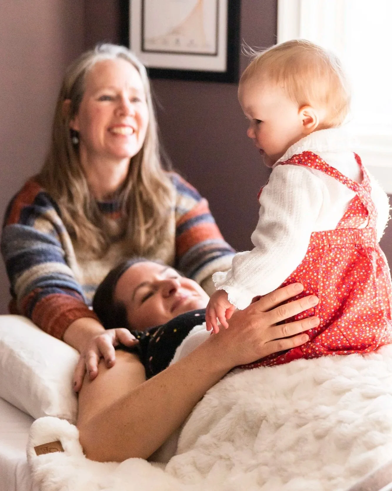A woman with long gray hair laughing while holding a young girl with red hair in a red dress with white dots. A child is lying on a bed with a white fluffy blanket in front of them. The setting appears to be a cozy room with a framed picture on the wall and a window with light streaming in.