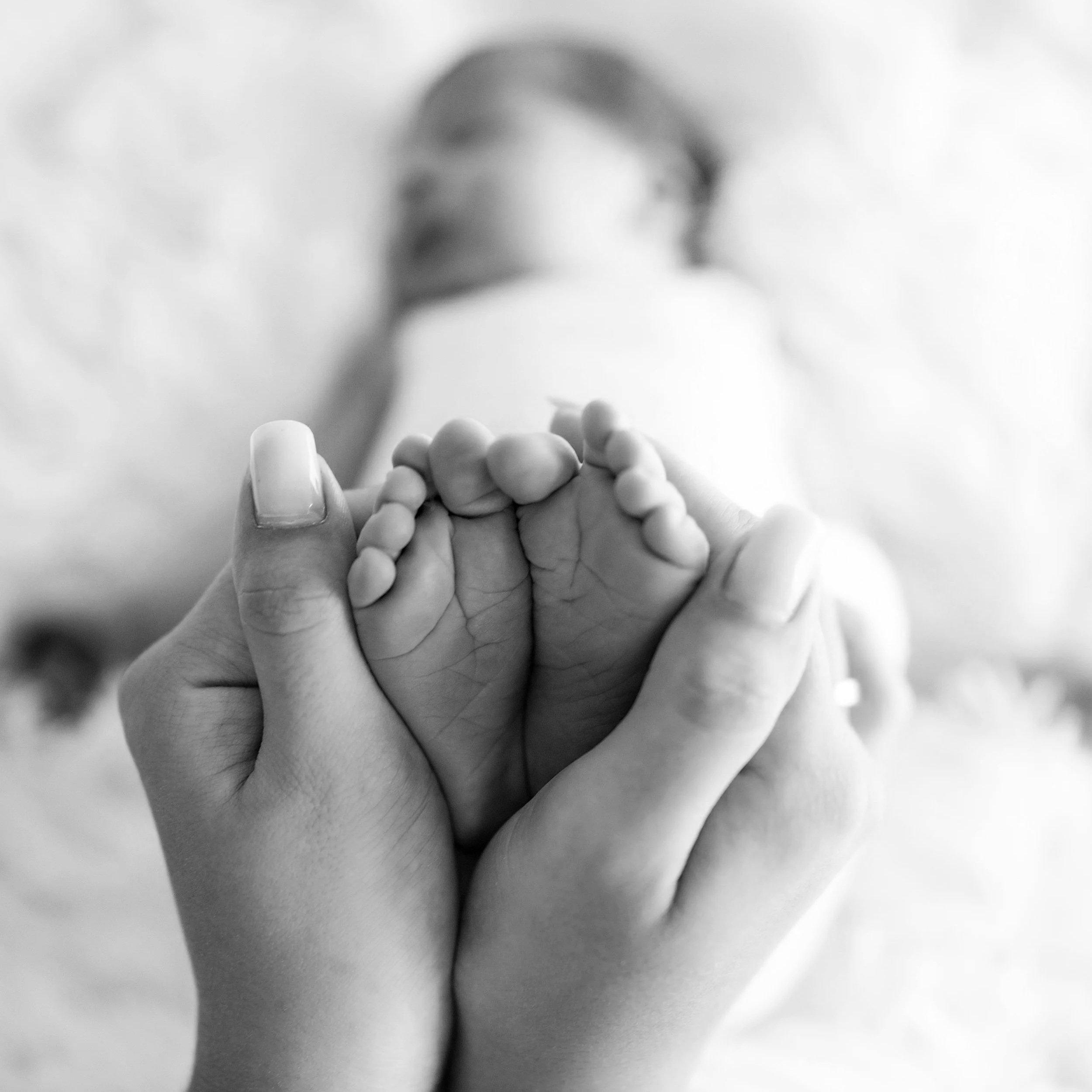 Close-up of a hand holding a tiny baby finger, with a blurred face of an adult in the background, in black and white.
