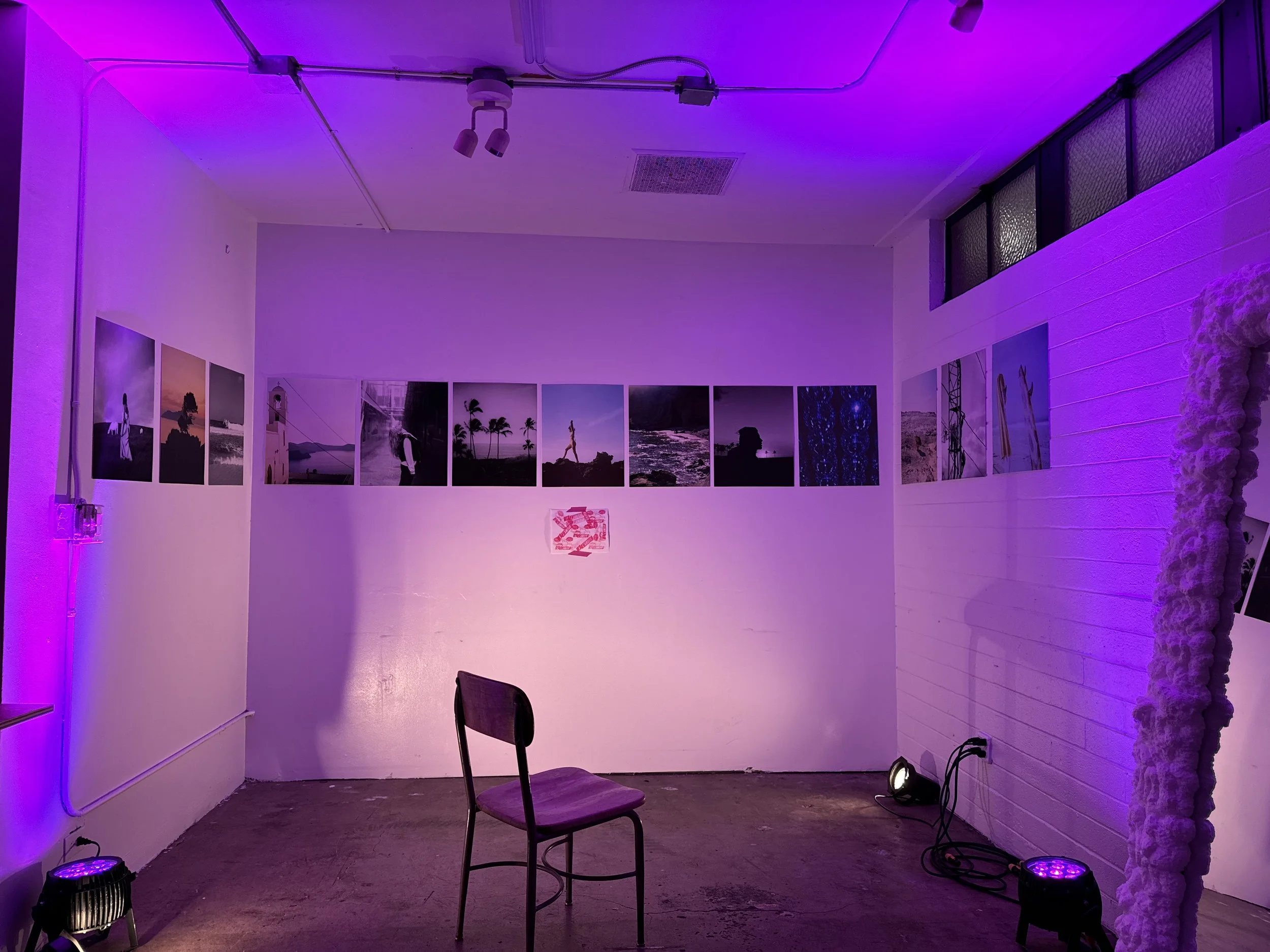Art gallery with purple lighting, displaying framed photographs on white walls, a wooden chair in the foreground, and decorative fabric on the right side.