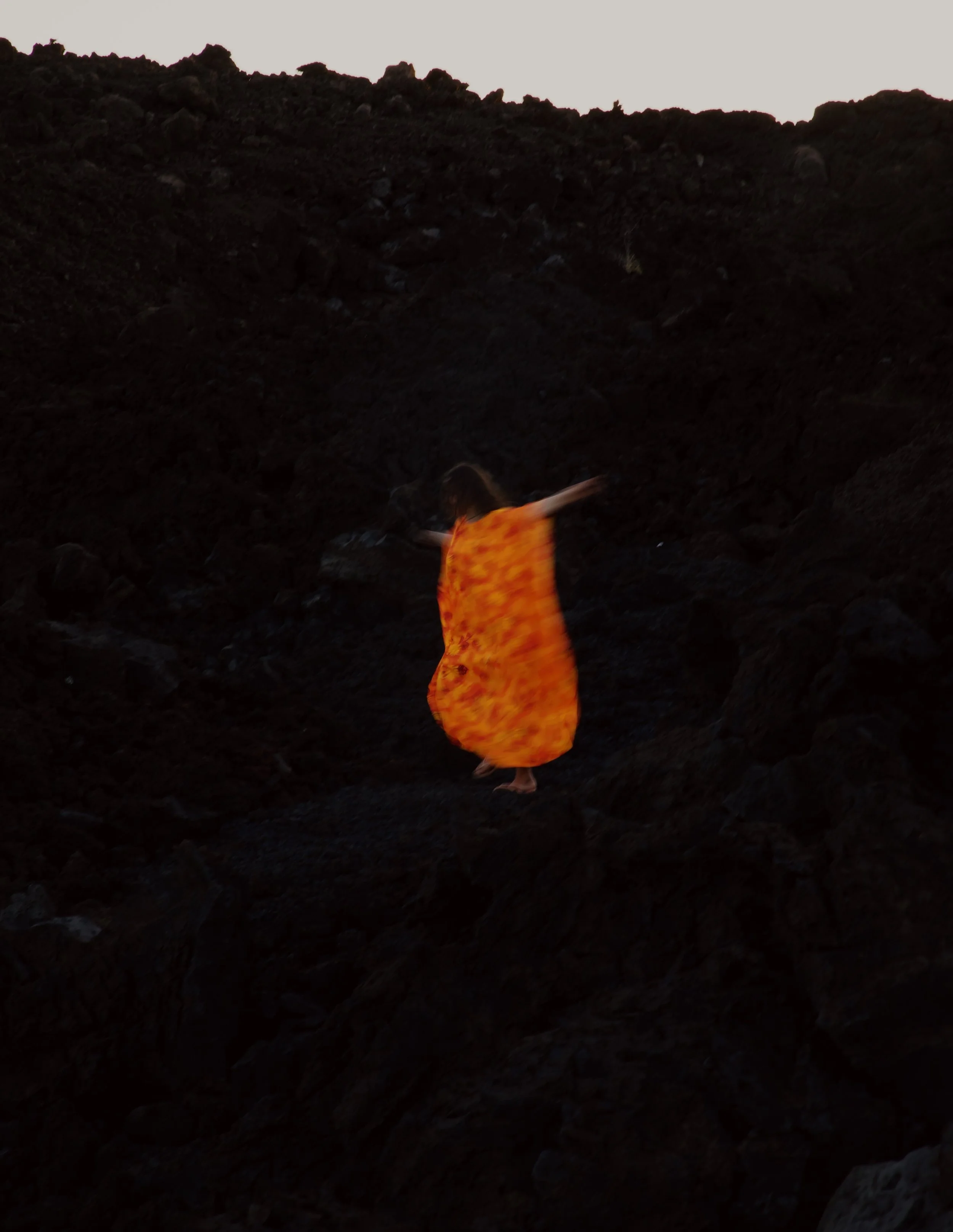 Child in an orange dress spinning in a dark rocky outdoor area during dusk.