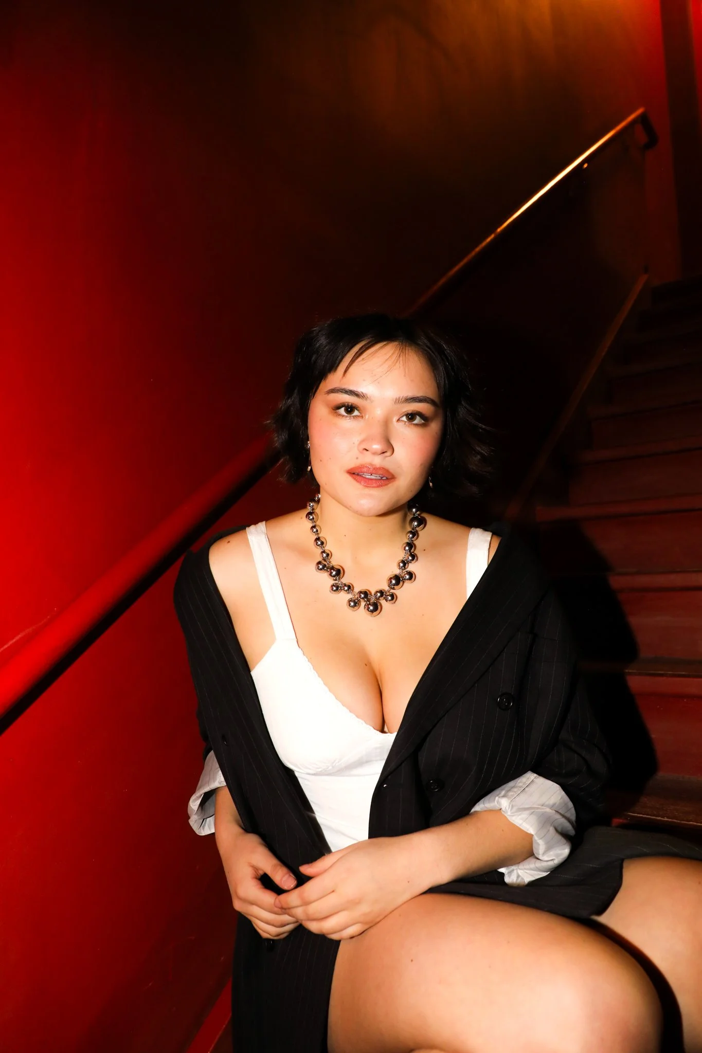 Young woman with black hair, wearing a black blazer draped over white top, silver necklace, and earrings, sitting on a staircase with a red wall background.