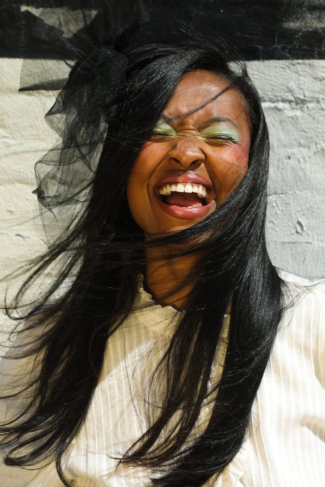 A woman with long black hair, wearing a large black tulle bow on her head, is laughing with her eyes closed in front of a white brick wall.